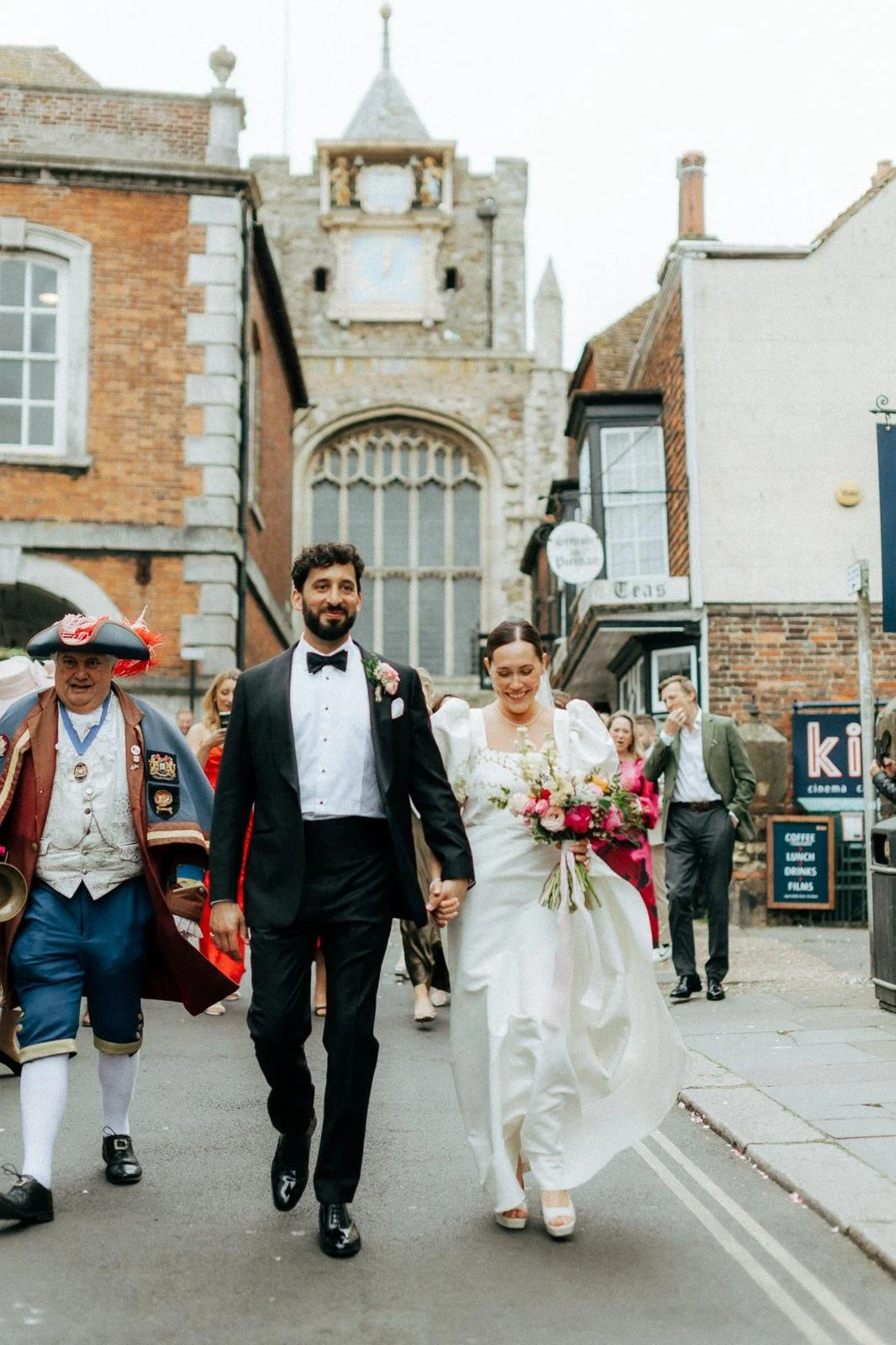 A newlywed couple walking hand-in-hand on a city street during their wedding day. The bride is carrying a bouquet of colorful flowers and wearing a white dress with puffed sleeves, while the groom is dressed in a black tuxedo with a bow tie. Several 