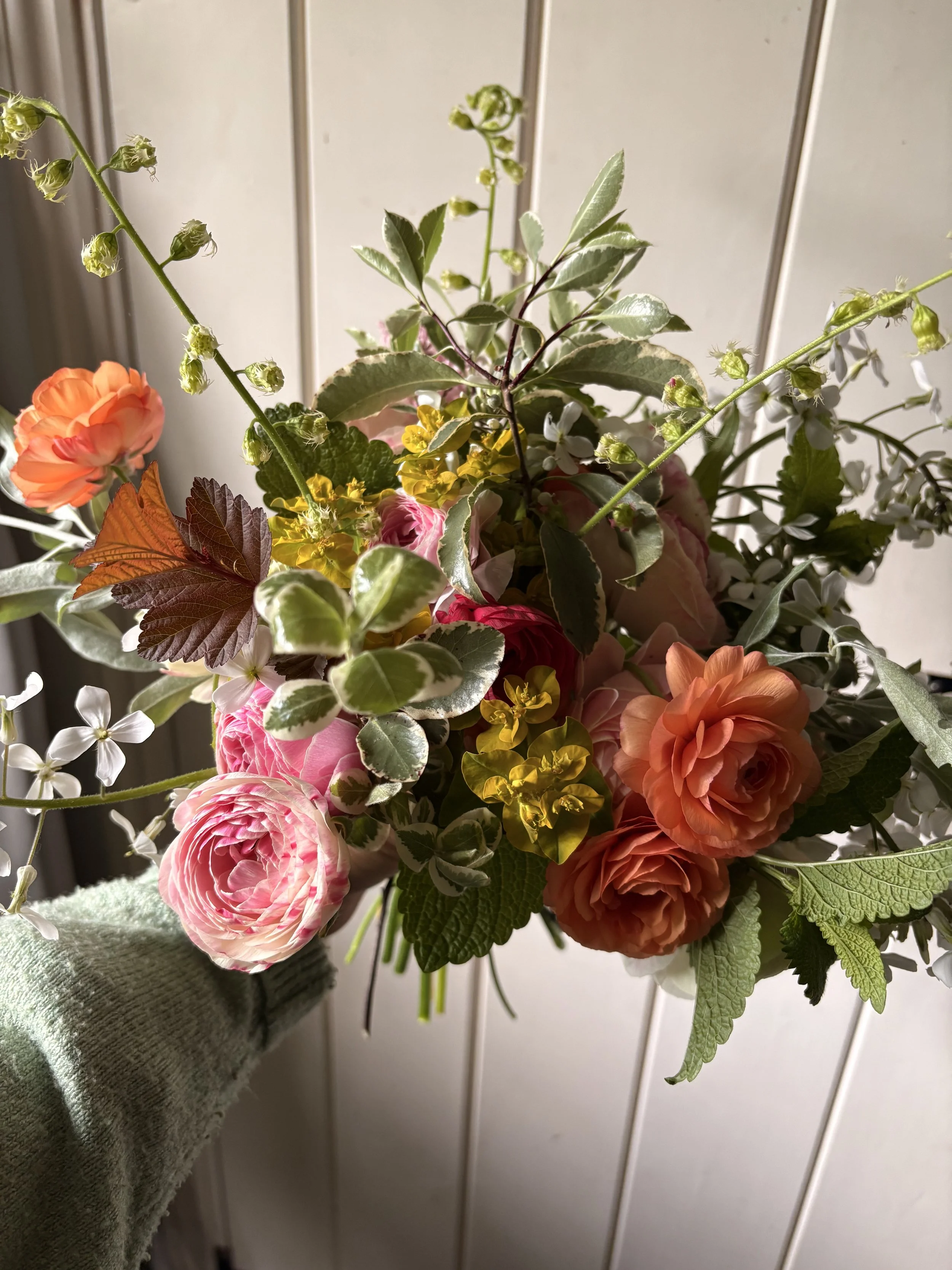 A colorful bouquet of flowers held by a person in a gray sleeve, featuring pink, orange, and yellow blooms, with green and variegated leaves, against a light-colored paneled wall.