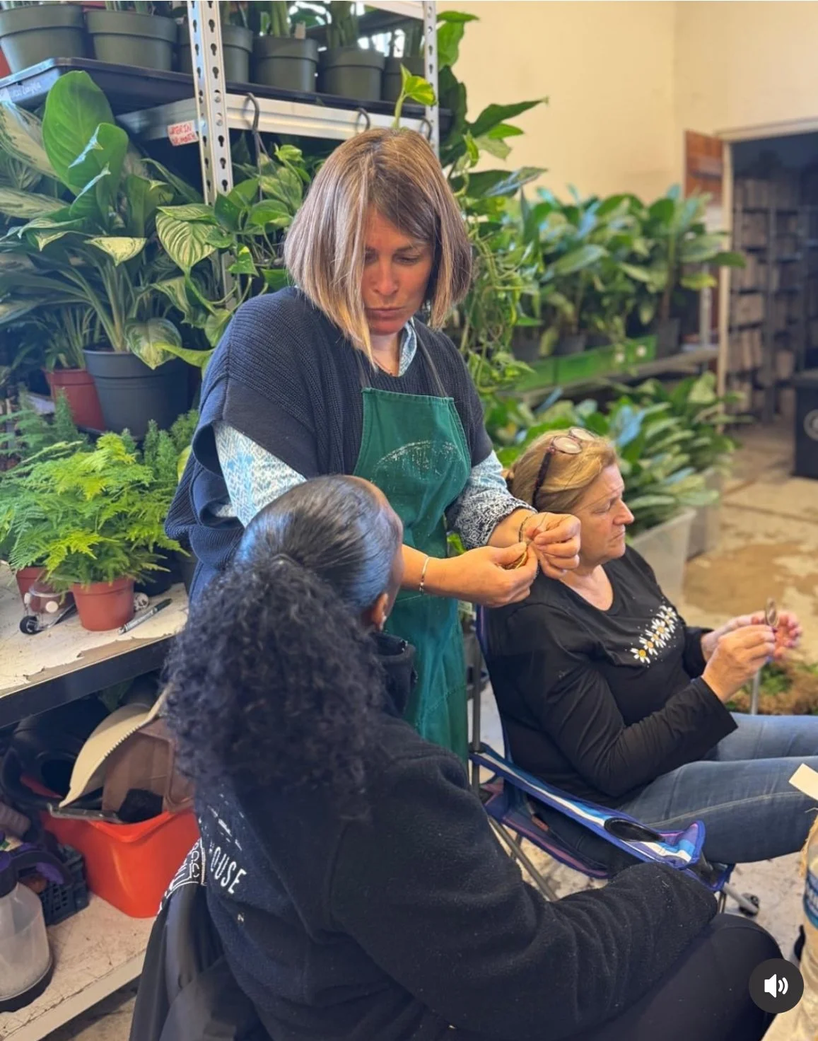 A woman is demonstrating a craft or plant-related activity to two women at a plant store or greenhouse, with shelves of potted plants in the background.