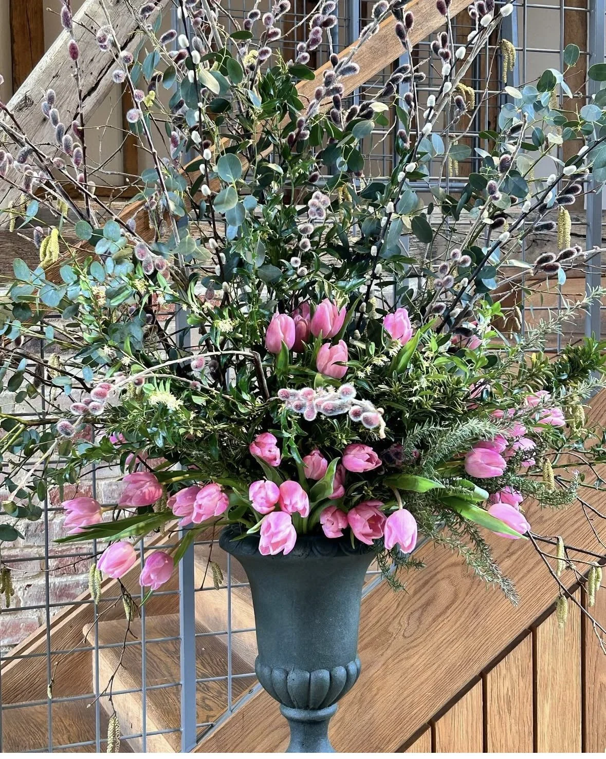 A large floral arrangement in a dark vase with pink tulips, greenery, and pussy willow branches on a wooden table against a brick wall and wire fence background.