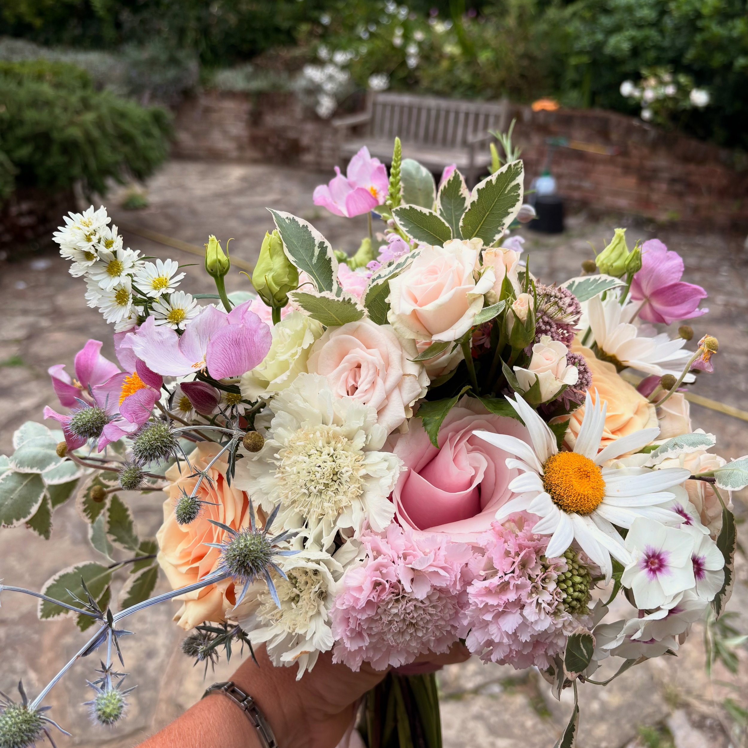 A hand holding a colorful bouquet of various flowers including roses, daisies, and carnations in an outdoor setting.
