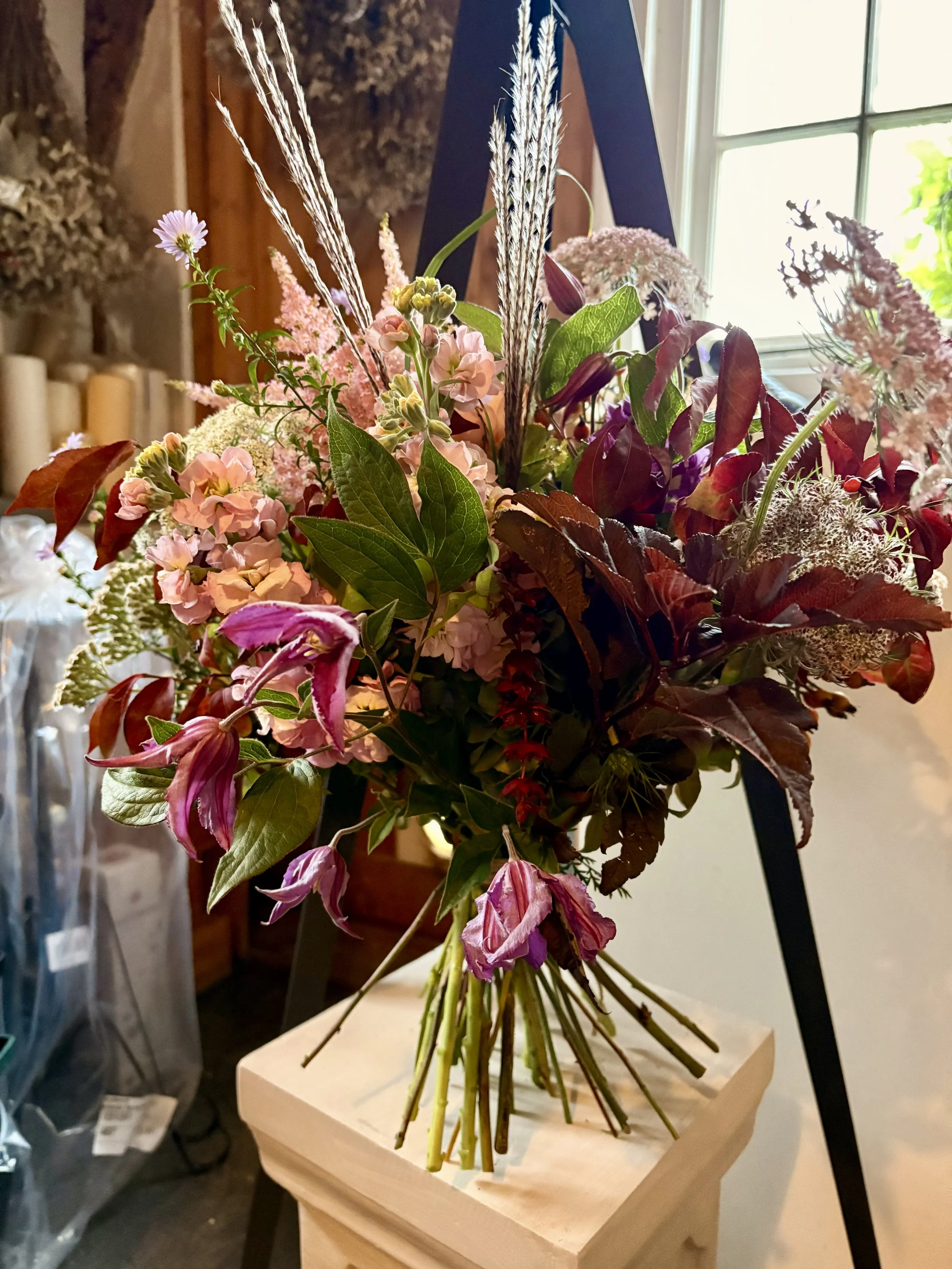 A colorful bouquet of flowers with pink, purple, and green hues arranged on a wooden pedestal near a window.