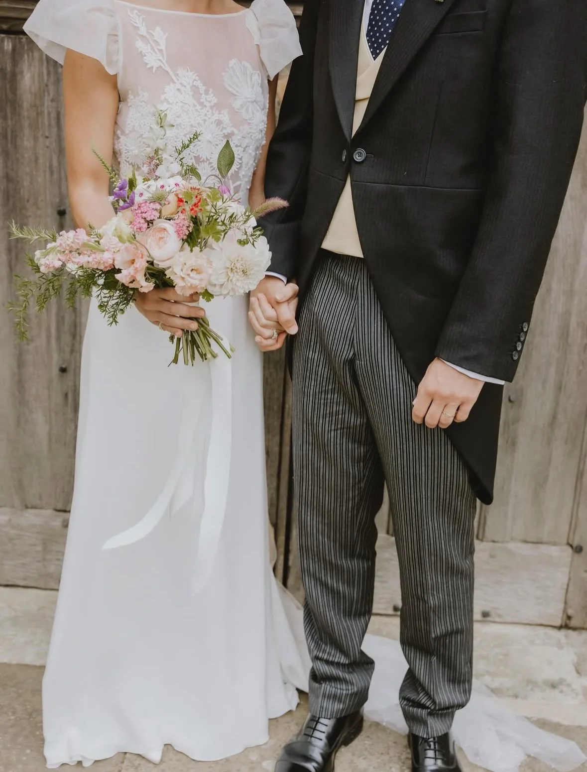 A bride in a white dress holding a bouquet, standing next to a groom in a black coat and striped pants, against a wooden backdrop.