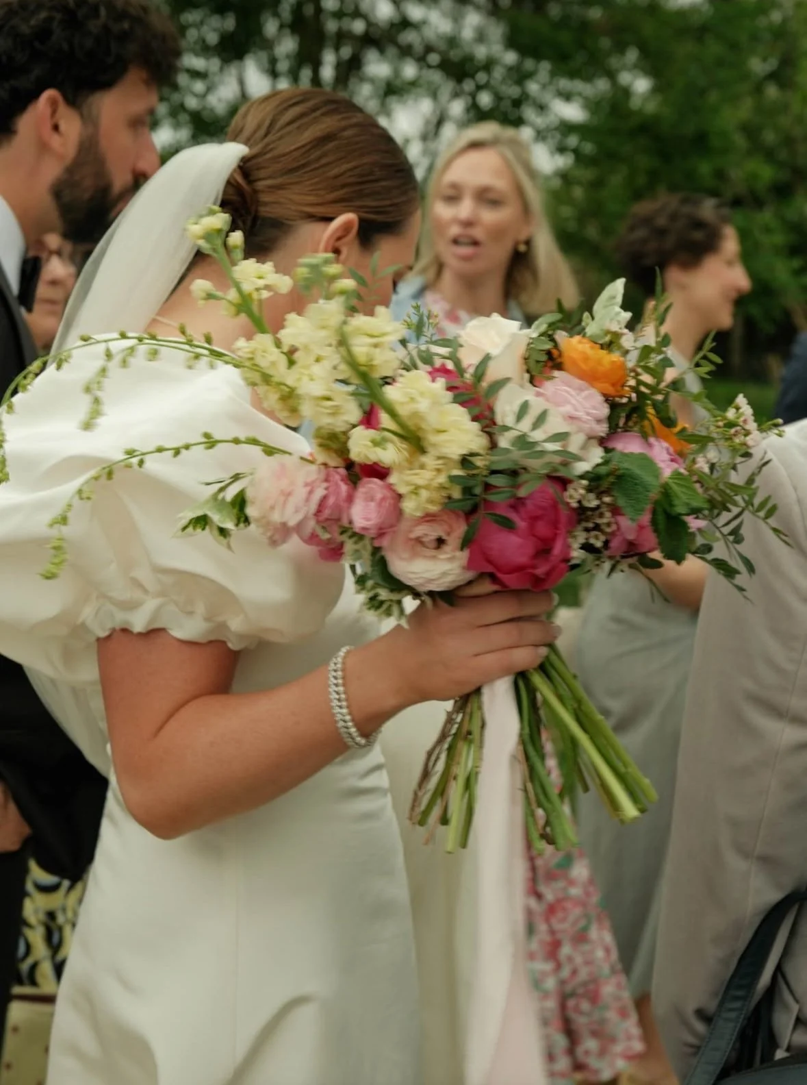 A bride holding a colorful bouquet of flowers, surrounded by wedding guests outdoors during a wedding ceremony.