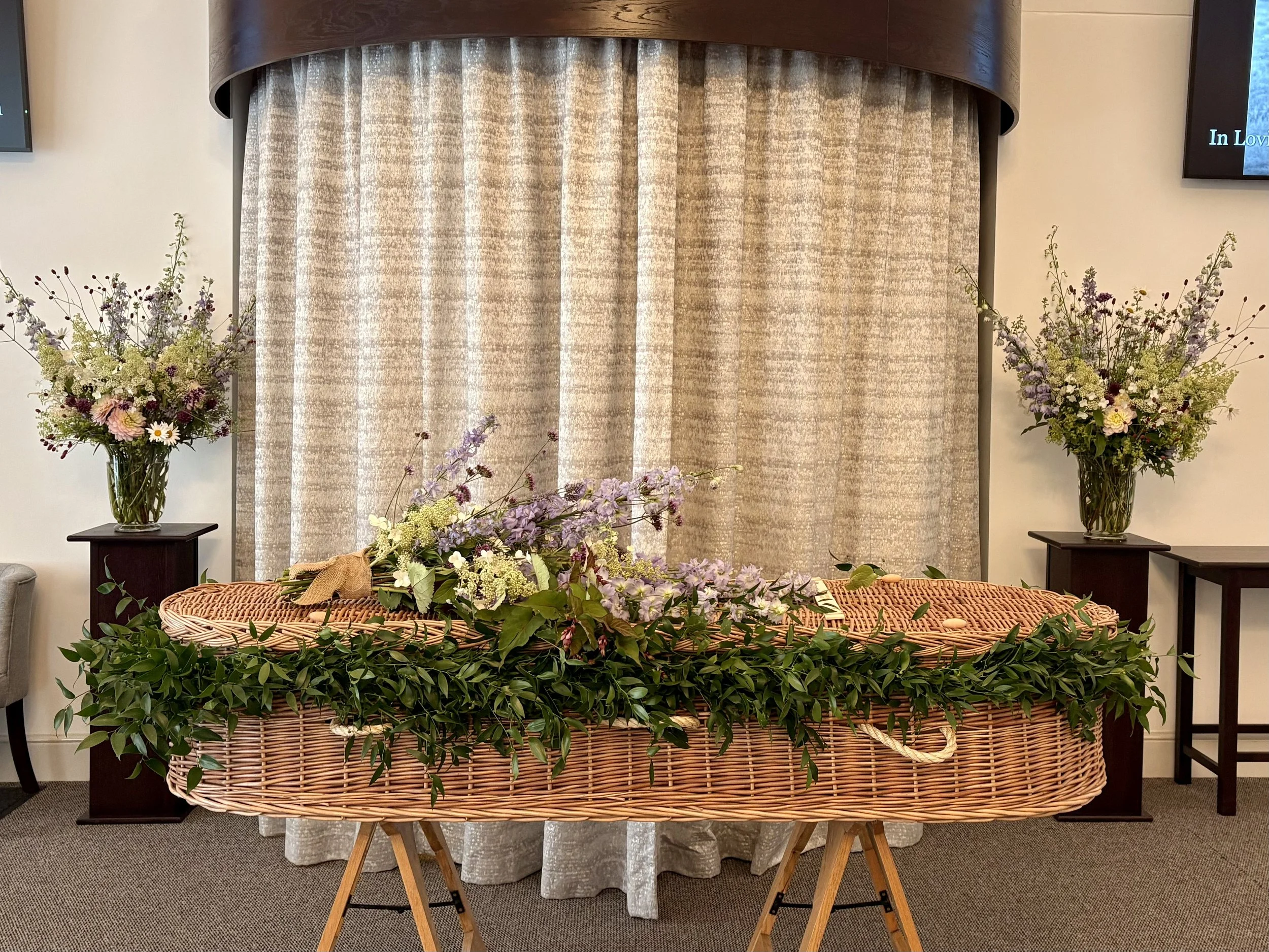 Wicker casket decorated with greenery and flowers, held by wooden stands, inside a room with beige curtains and floral arrangements on dark wood tables.