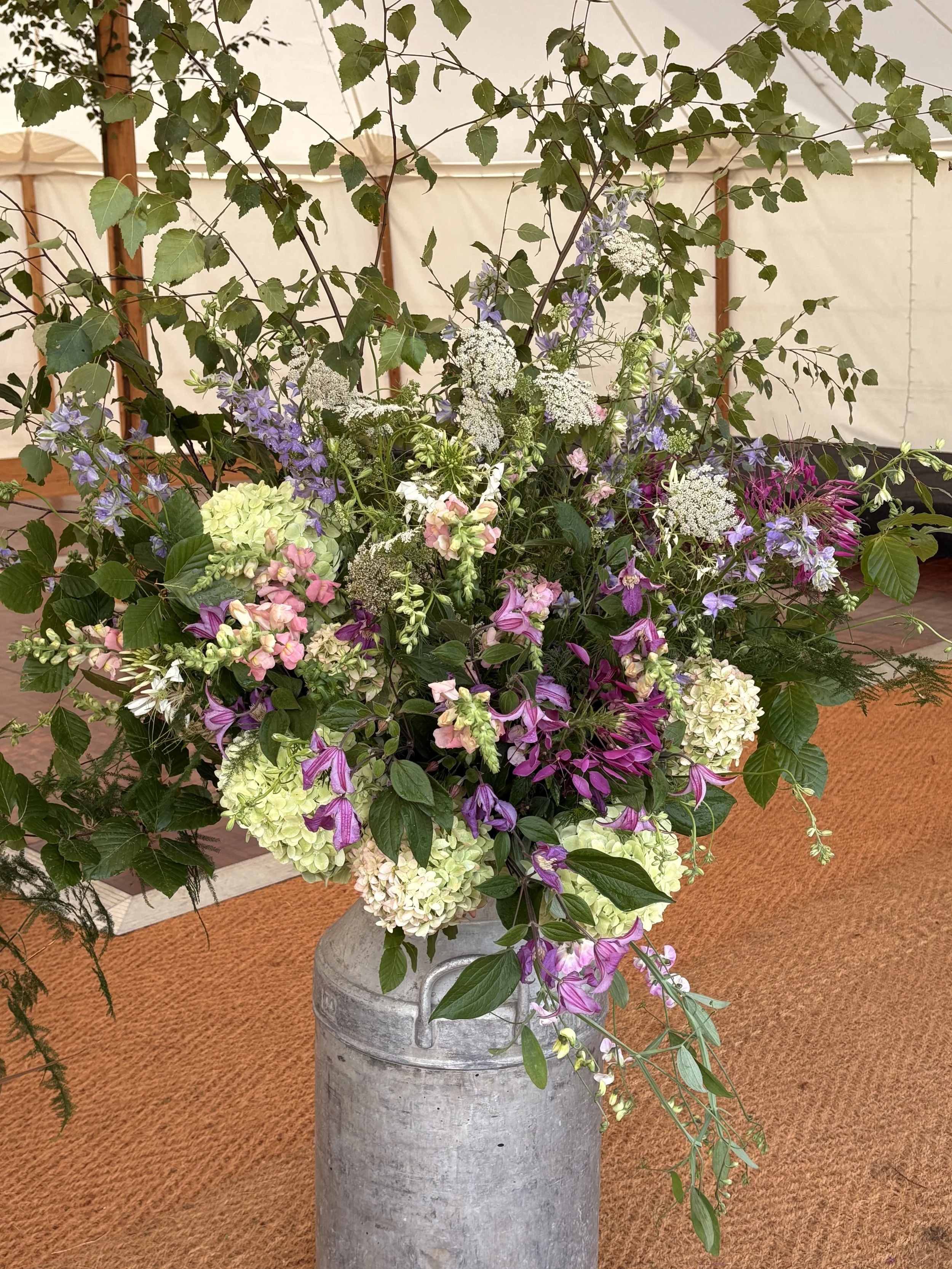 A large bouquet of mixed flowers in a rustic metal milk can, set on a tan textured floor inside a tent or pavilion.