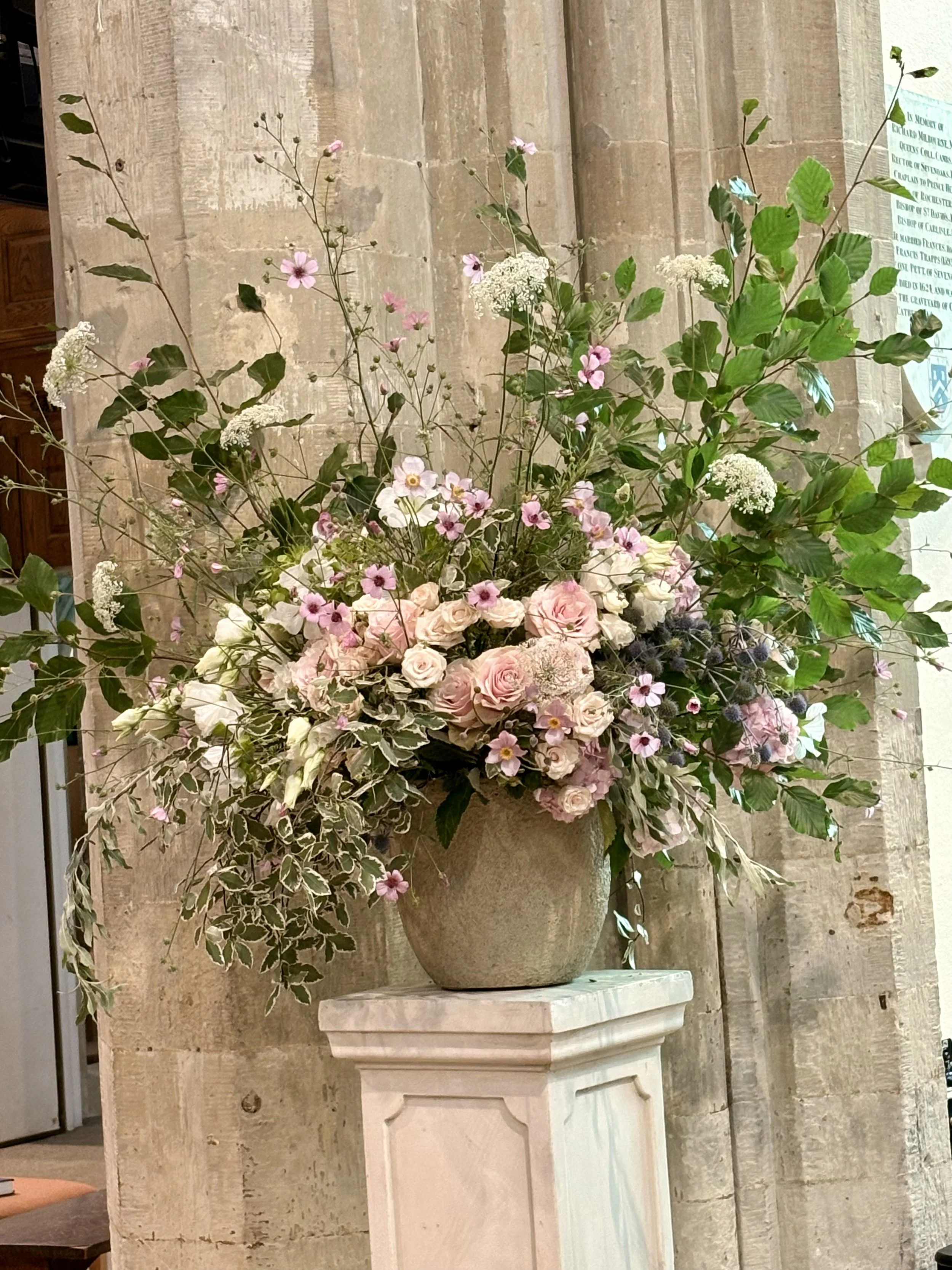 A large floral arrangement in a gray vase on a white pedestal, with pink, white, and green flowers and foliage, set against a stone wall.