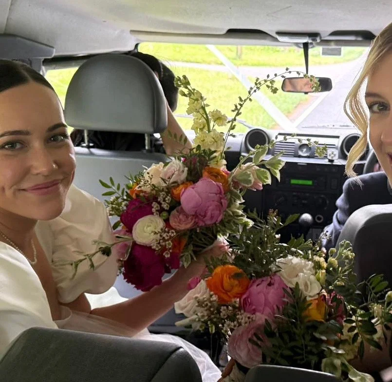 A bride and her bridesmaid sitting in the front seats of a car holding large bouquets of colorful flowers.