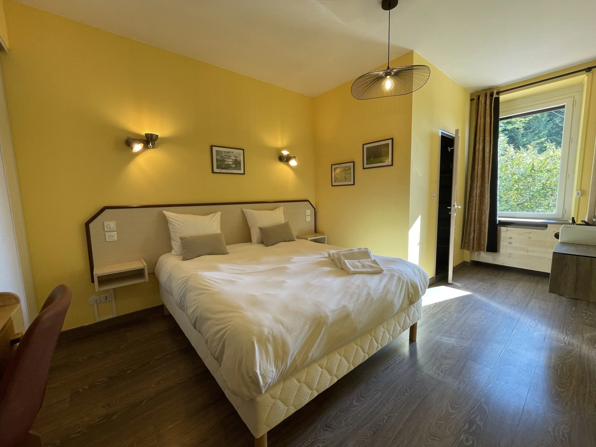 Chambre d'hôtel avec lit double, murs jaunes, fenêtre ouverte laissant entrer la lumière et vue sur la nature, décor simple et lumineux.