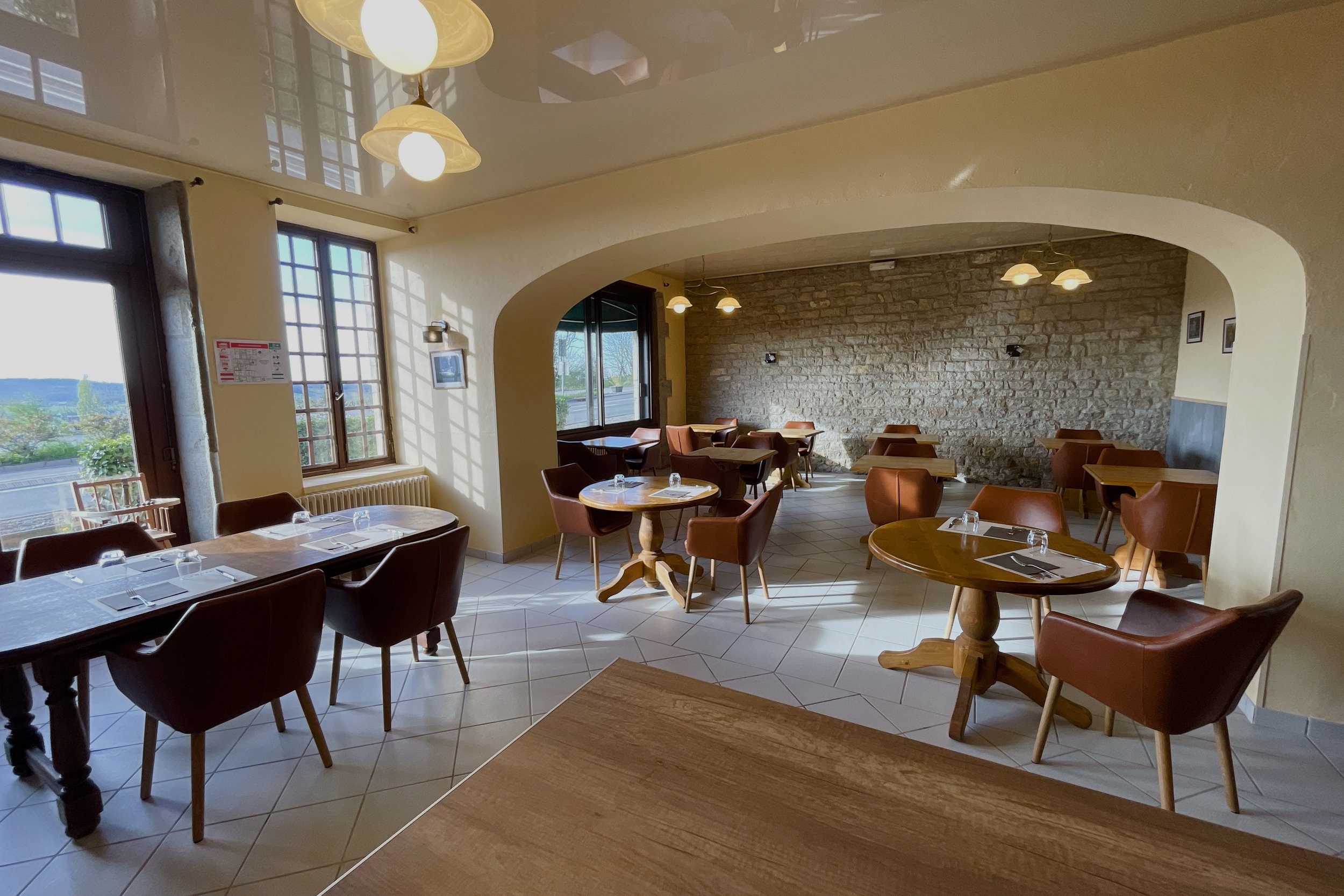 Salle de restaurant lumineuse avec tables en bois, chaises marron, mur en pierre apparente et grandes fenêtres donnant sur l'extérieur.
