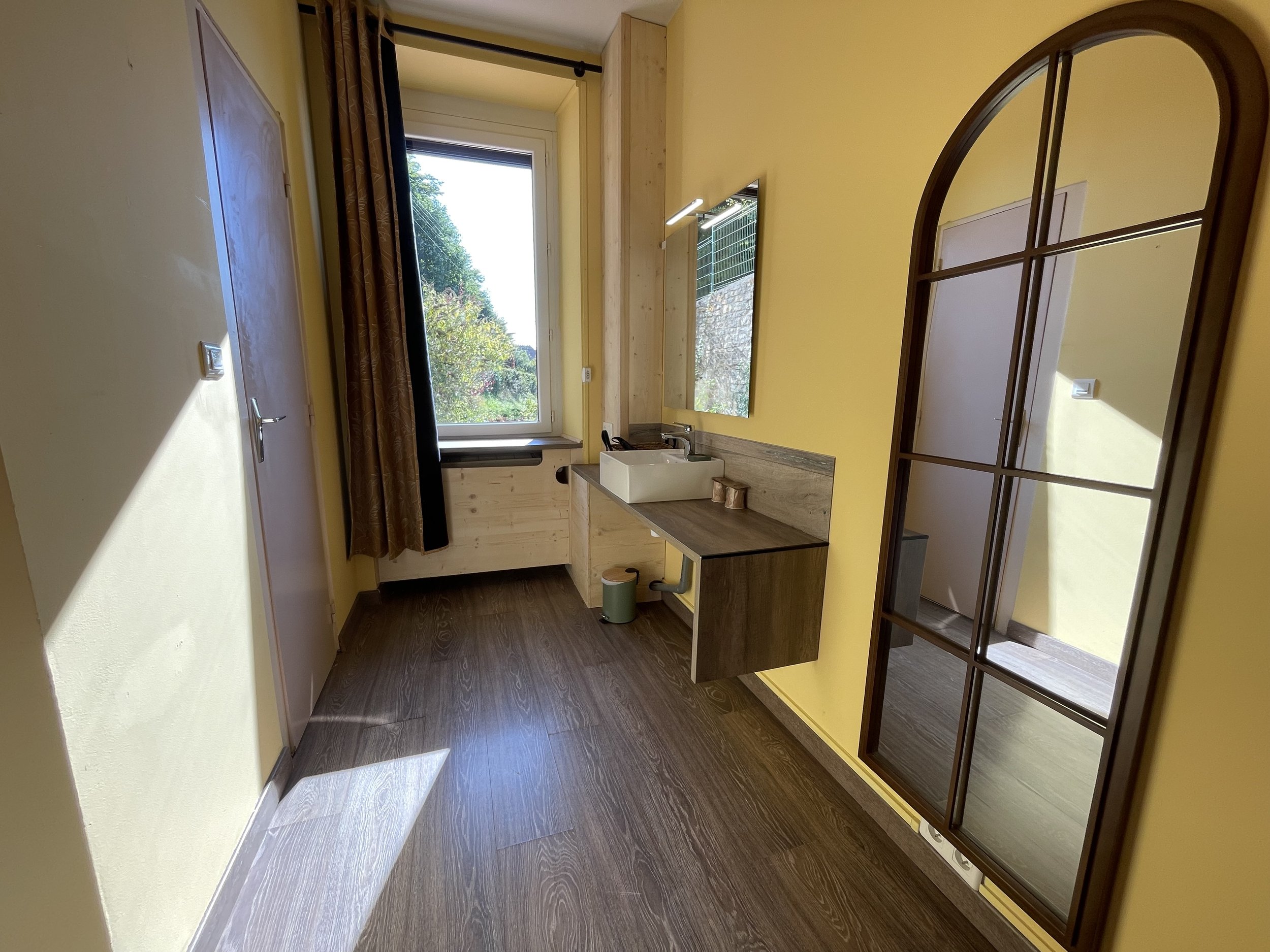 Intérieur d'une pièce avec murs jaunes, sol en bois foncé, une fenêtre laissant passer la lumière, un miroir en miroir noir, un évier blanc sur un meuble en bois, et un rideau marron près de la fenêtre.