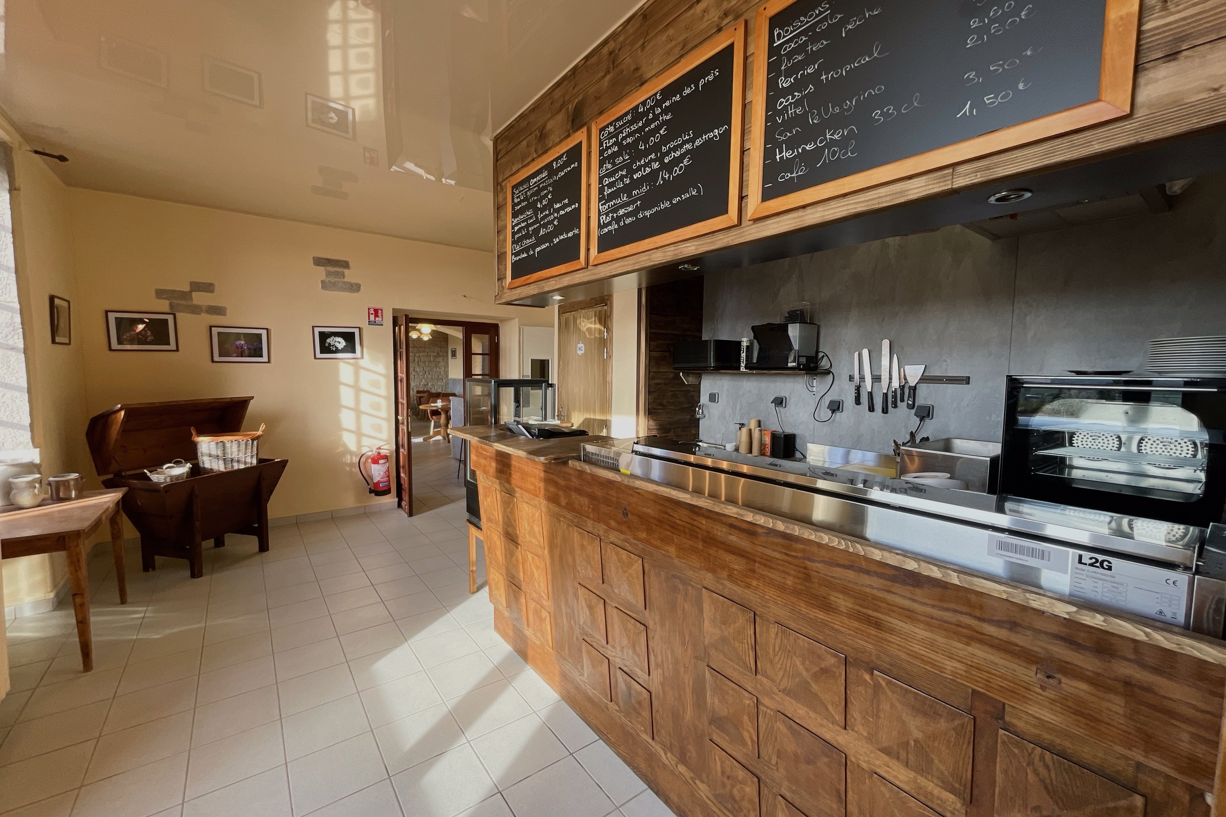 Intérieur d'un café avec un comptoir en bois, des tableaux de menus au-dessus, et des tables en arrière-plan. Un four, des couteaux sur un support mural, et une corbeille sur un meuble à gauche sont visibles. Il y a aussi une porte ouverte menant à u