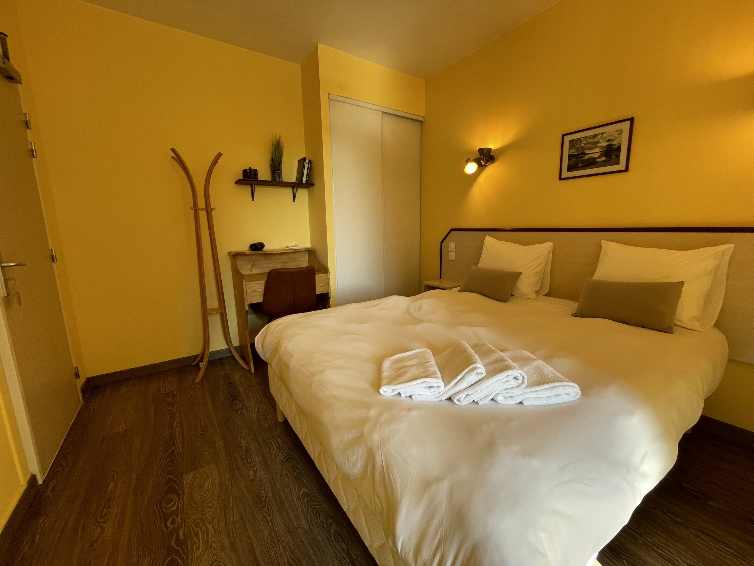 Chambre d'hôtel avec un lit double, des serviettes blanches pliées sur le lit, et un mur jaune avec une lampe et une photo encadrée.