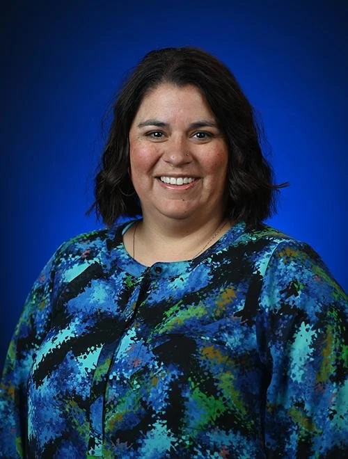 A woman with shoulder-length brown hair smiling, wearing a patterned blouse, against a blue background.