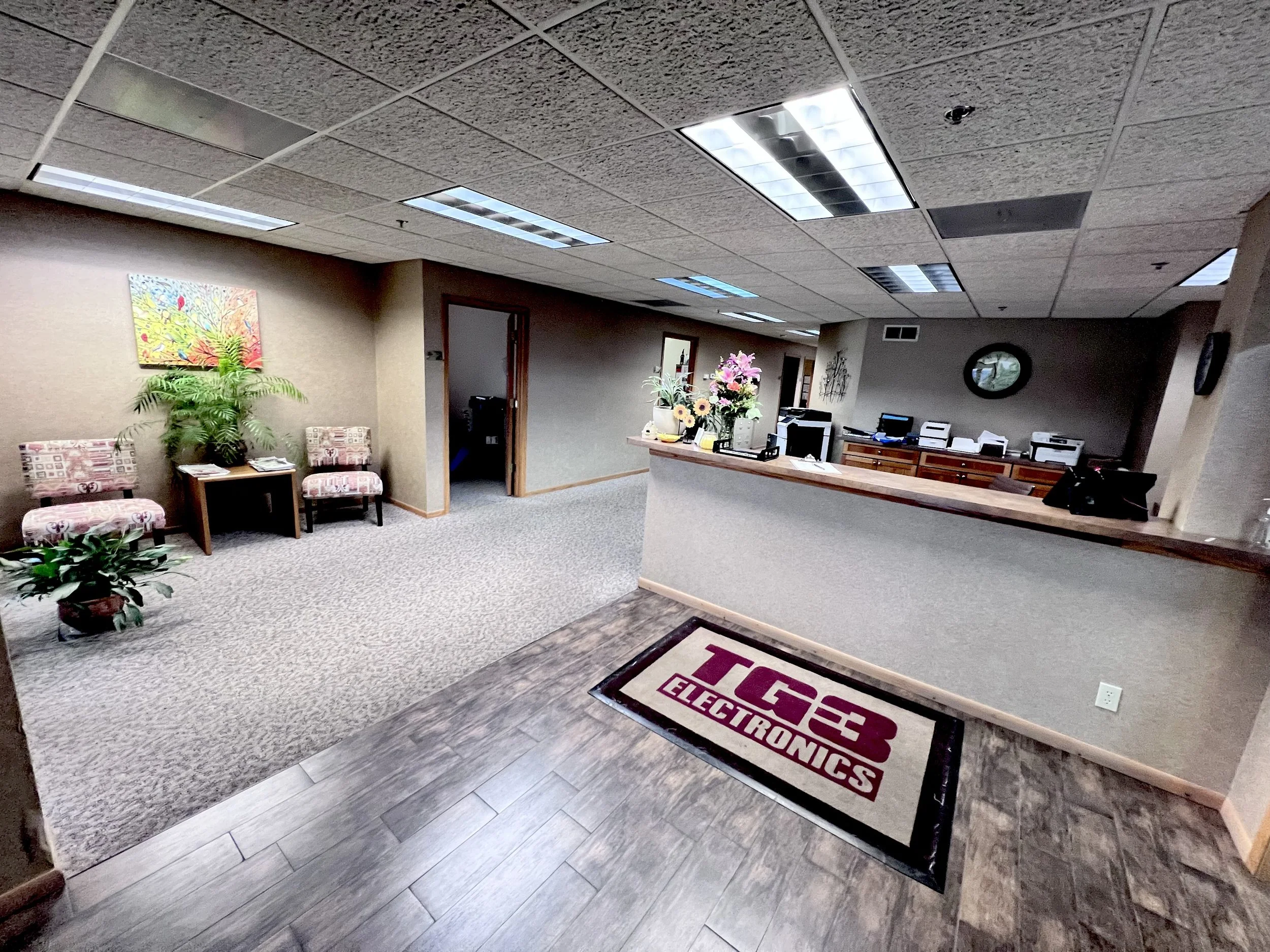 Interior view of an electronics store or office reception area with a greeting mat, reception desk with flowers, bulletin boards, and waiting area with chairs and plants.
