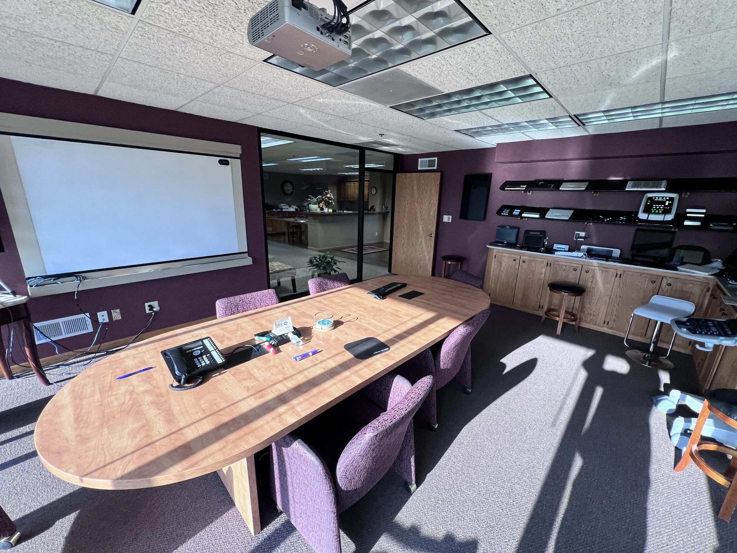 Conference room with a large wooden oval table, purple chairs, a whiteboard, and various office equipment, with sunlight casting shadows on the carpeted floor.
