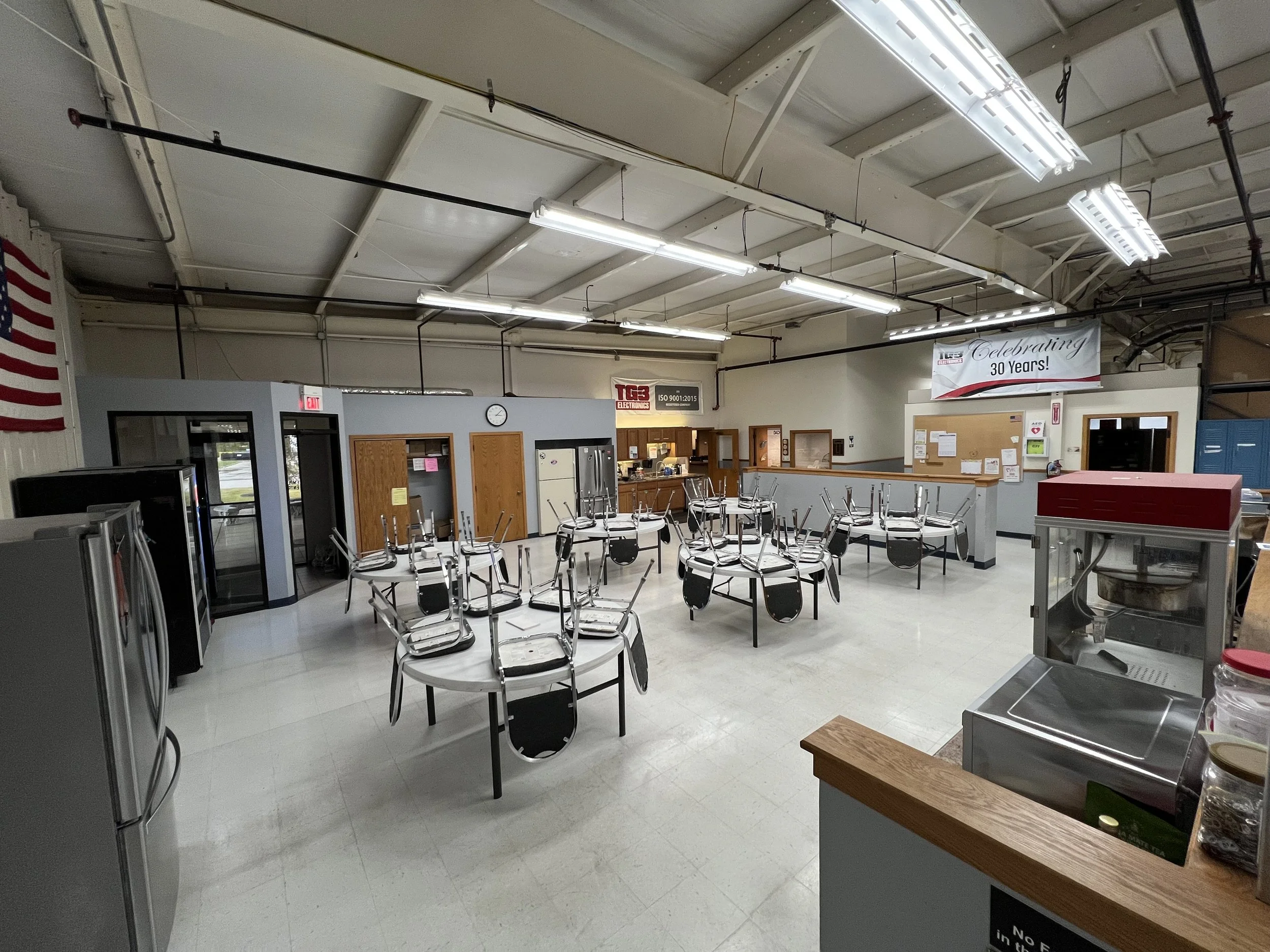 Empty cafeteria or break room with chairs stacked on tables, vending machines, refrigerator, and bulletin boards. Fluorescent lighting and a banner celebrating 30 years.