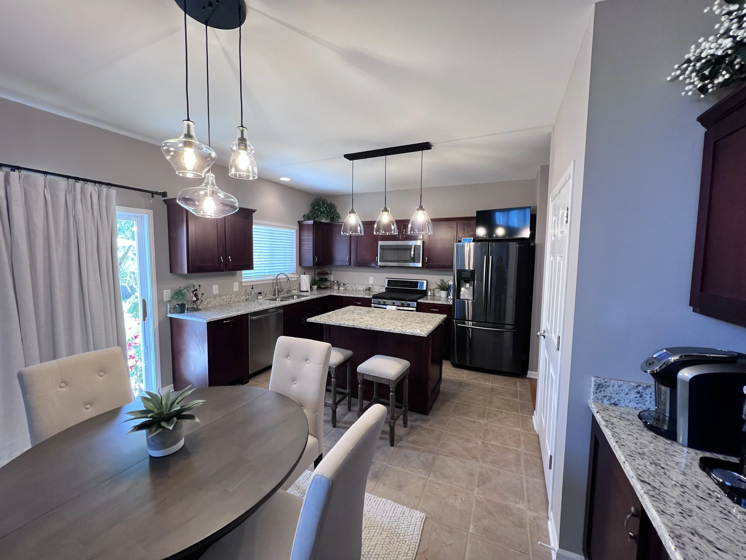 Modern kitchen with dark wood cabinets, granite countertops, stainless steel appliances, pendant lighting over the island, and a round dining table with beige chairs.