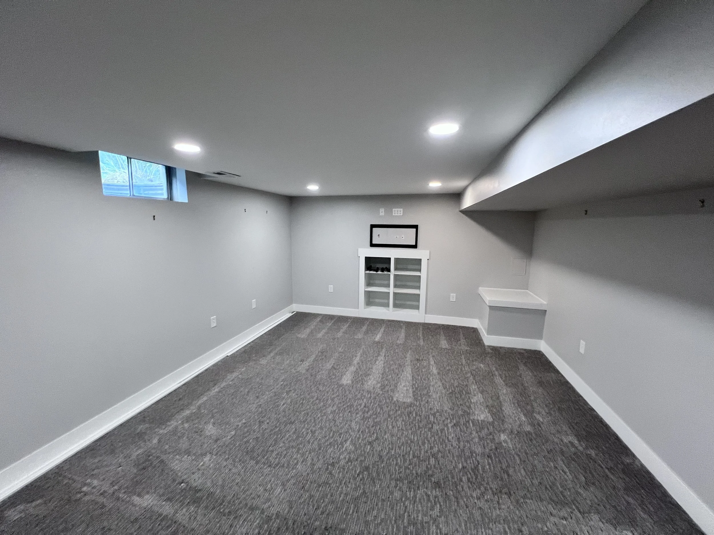 Empty finished basement with gray walls, gray carpet, recessed lighting, small window, and built-in shelving.