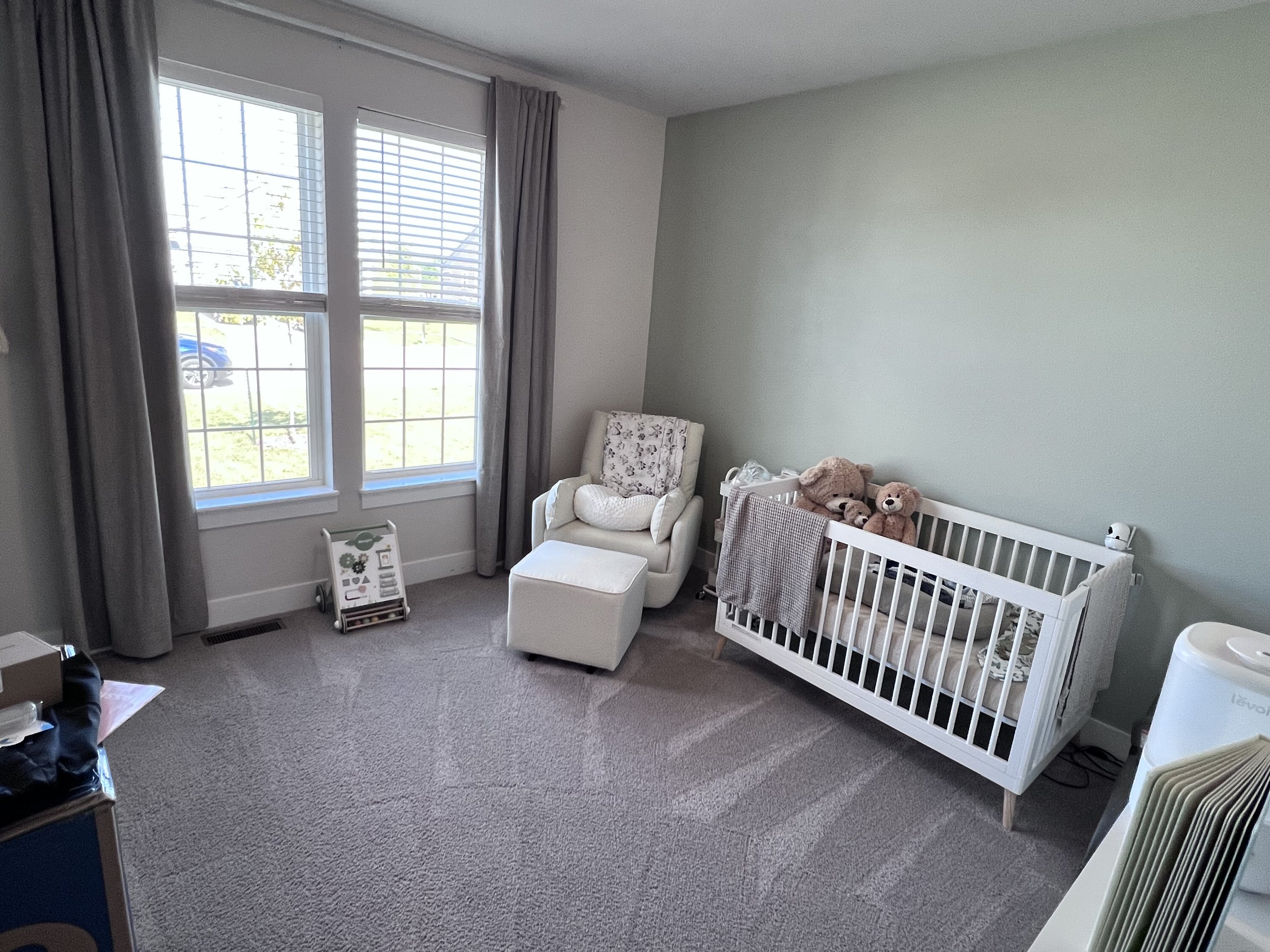 A nursery with large windows, gray curtains, a white armchair with a matching ottoman, a crib with teddy bears, and a small white side table.