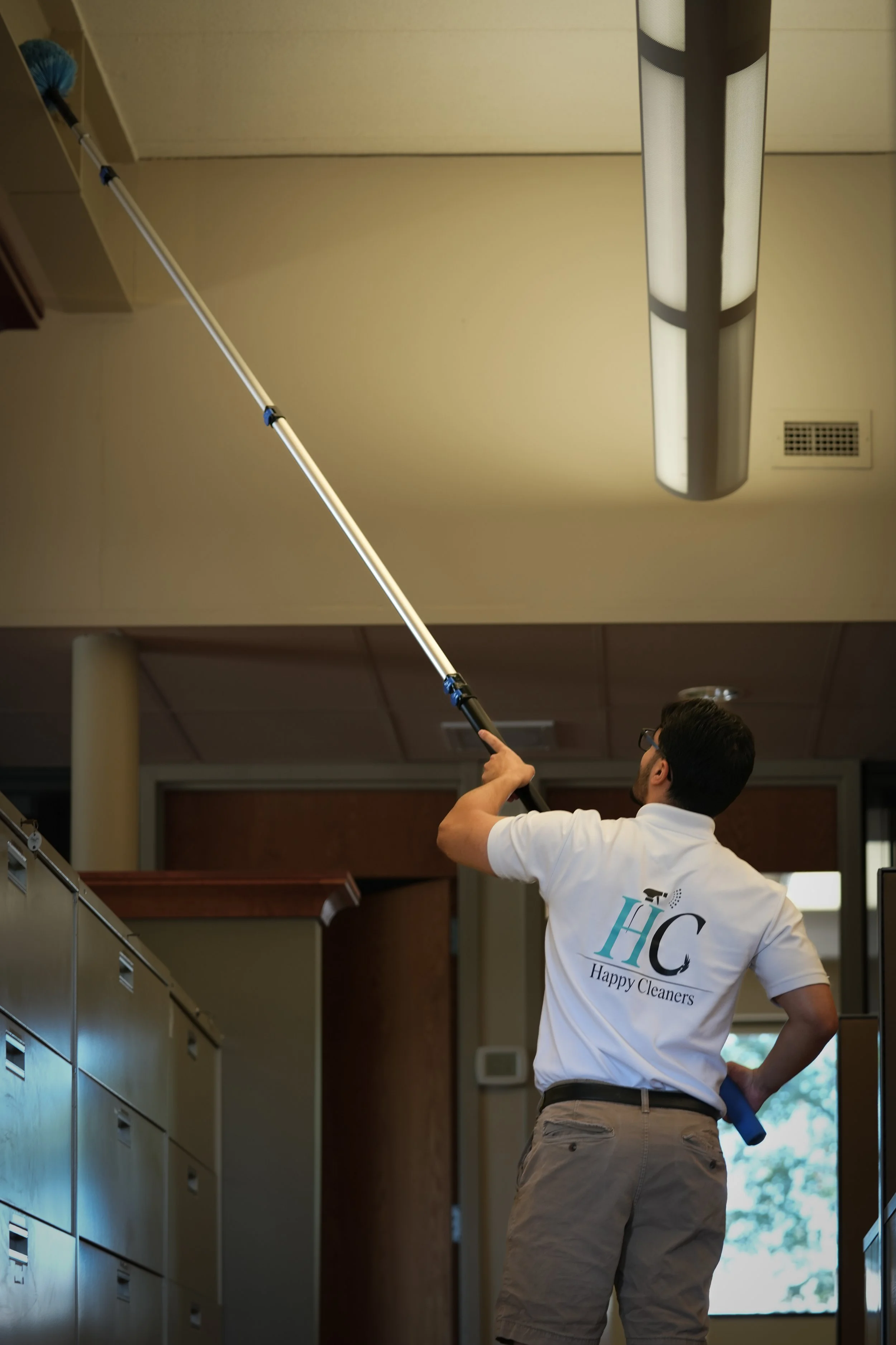 A man using a long-handled cleaning tool to clean a ceiling vent or light fixture, wearing a white polo shirt with the logo 'HC Happy Cleaners'.