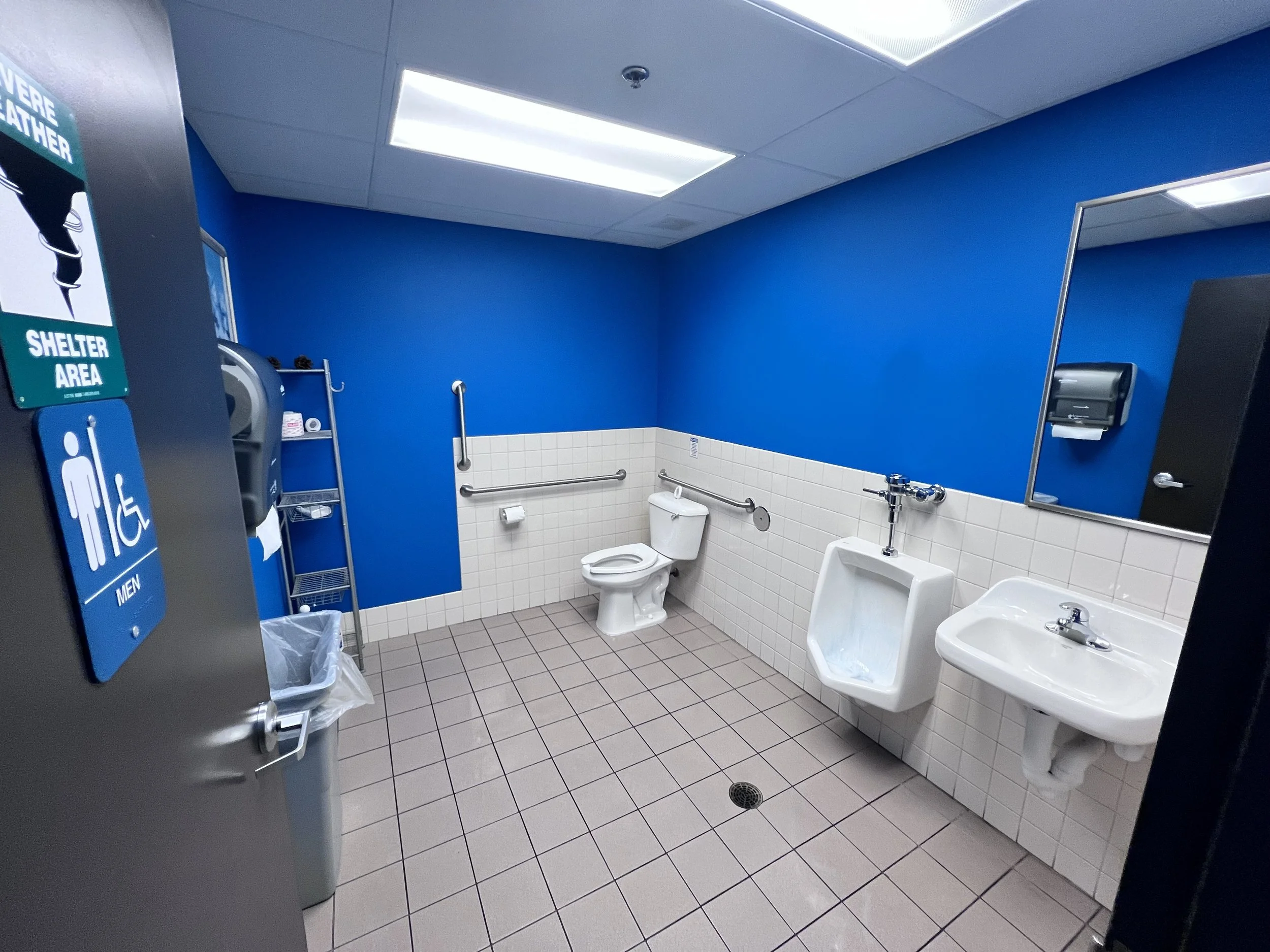 Public men's restroom with blue painted walls, white tiles, a toilet, urinal, sink, and accessibility features including handrails and a non-slip floor.
