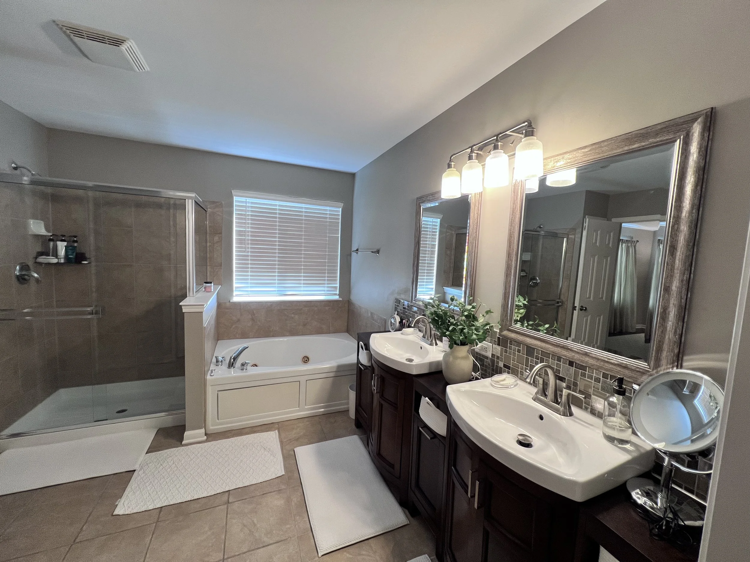 A spacious bathroom with a glass-enclosed shower, a bathtub, and two sinks with mirrors and lights above. There are white towels, a potted plant, and toiletries on the counters. A large window with blinds lets in natural light.