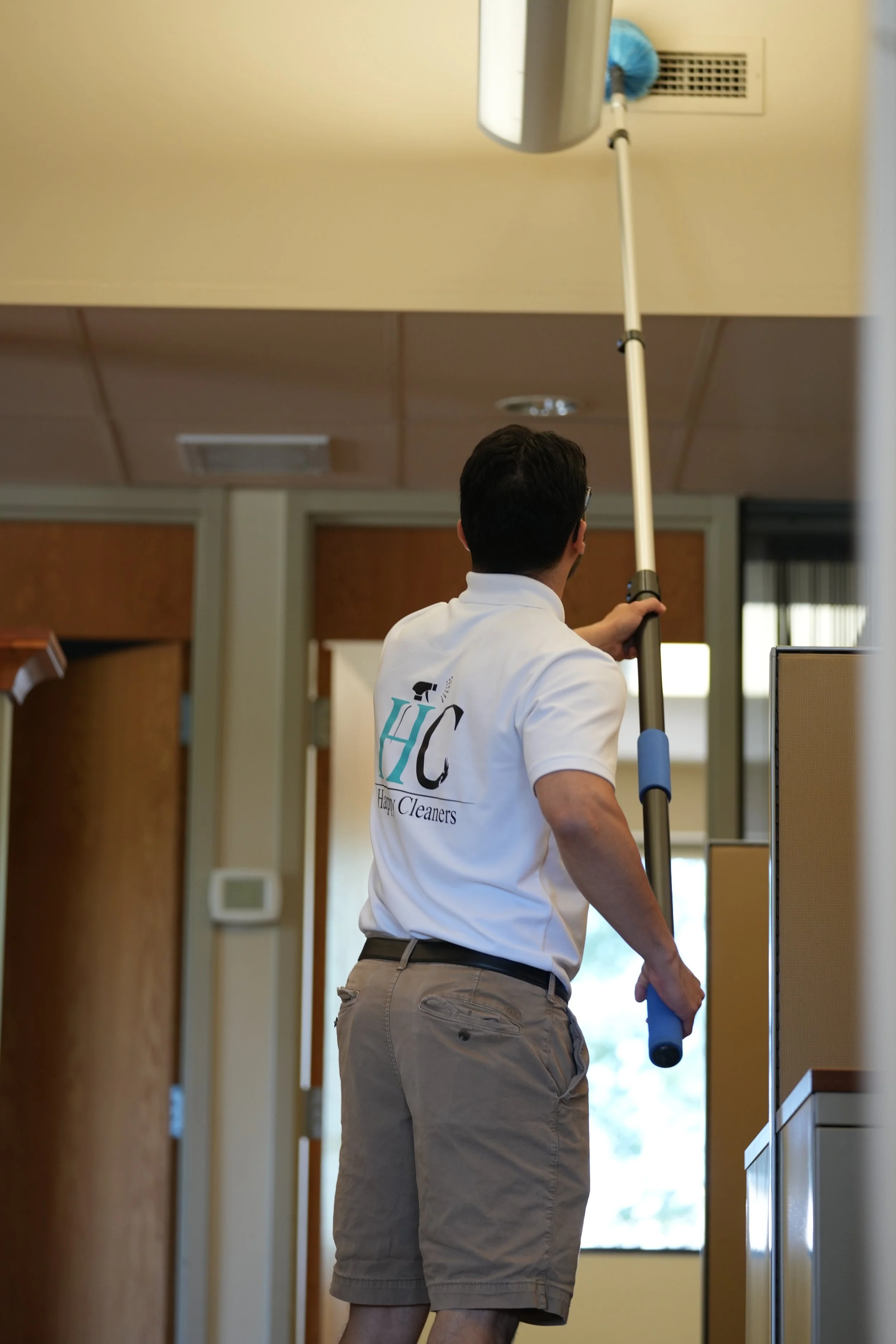 A person wearing a company shirt cleaning a ceiling vent with an extendable duster.