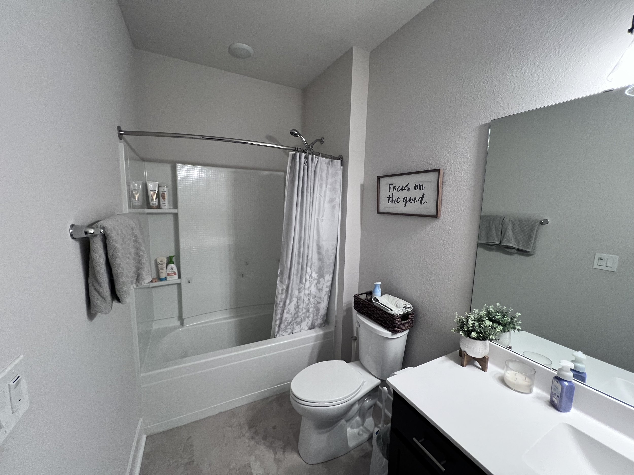 Bathroom with bathtub and shower, gray towels, toilet, white countertop with toiletries, large mirror, wall art saying 'Focus on the good,' and a potted plant.