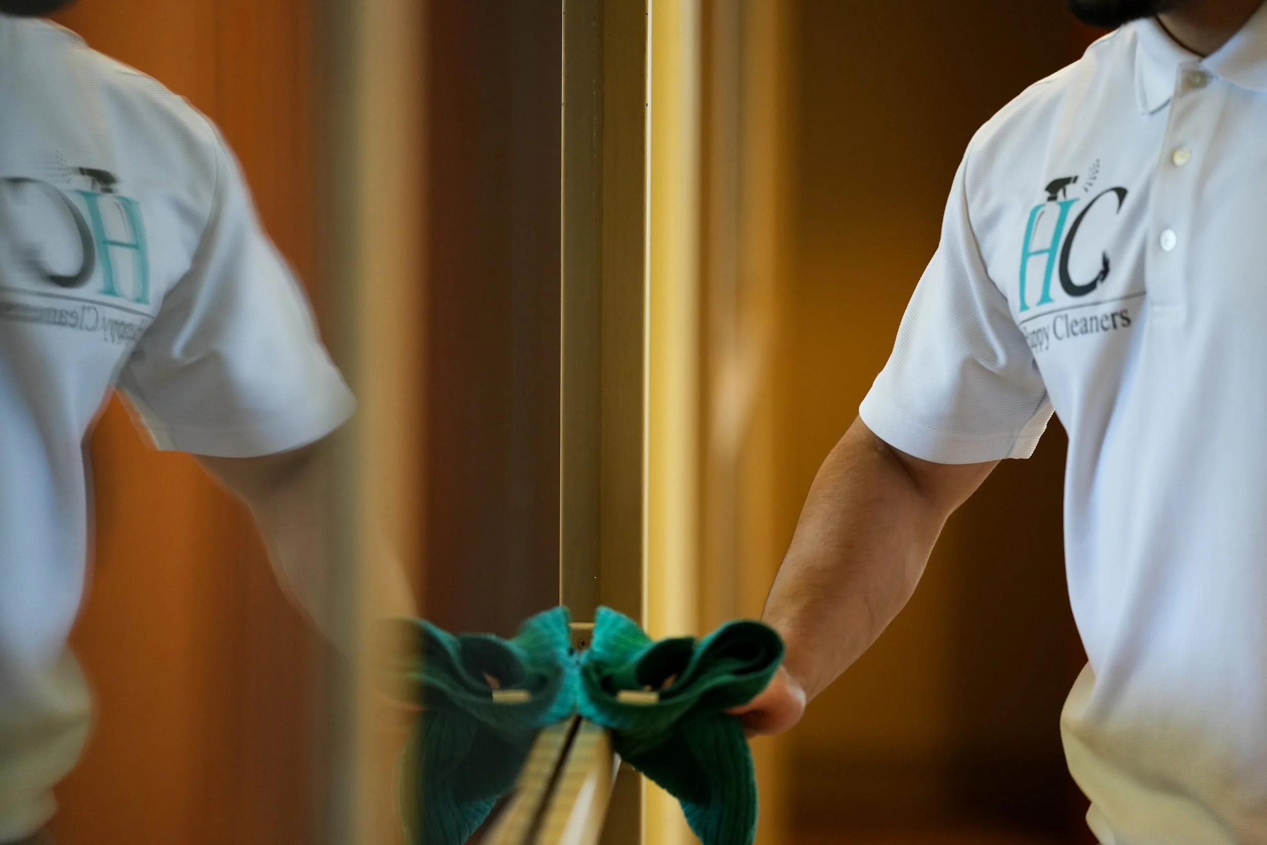 Person cleaning a mirror with a cloth, wearing a white polo shirt with a company logo.
