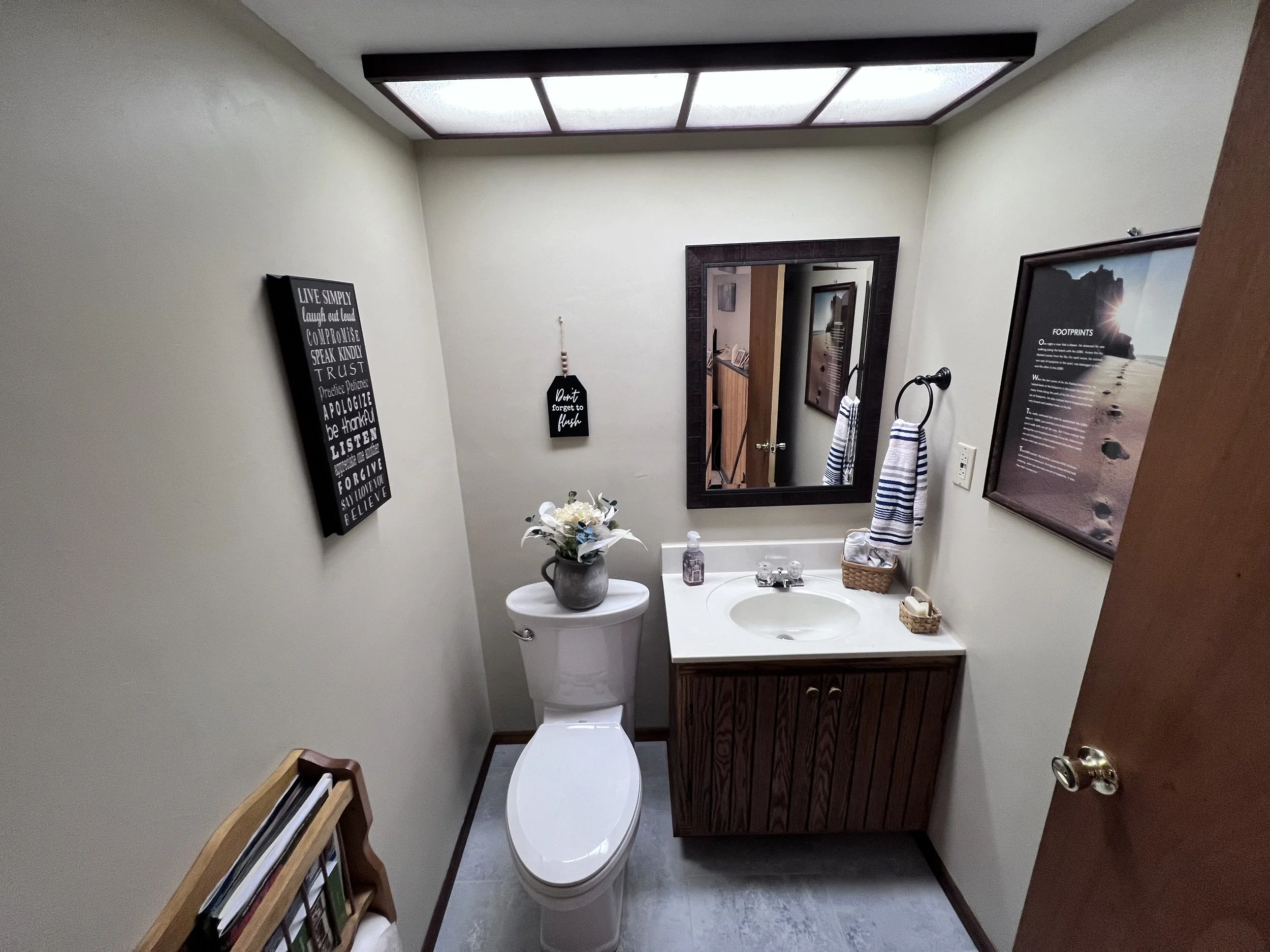 Small bathroom with a white toilet, a wooden vanity with a white countertop and sink, a mirror on the wall, framed artwork, a small dark wall hanging with text, towels, and decorative items.