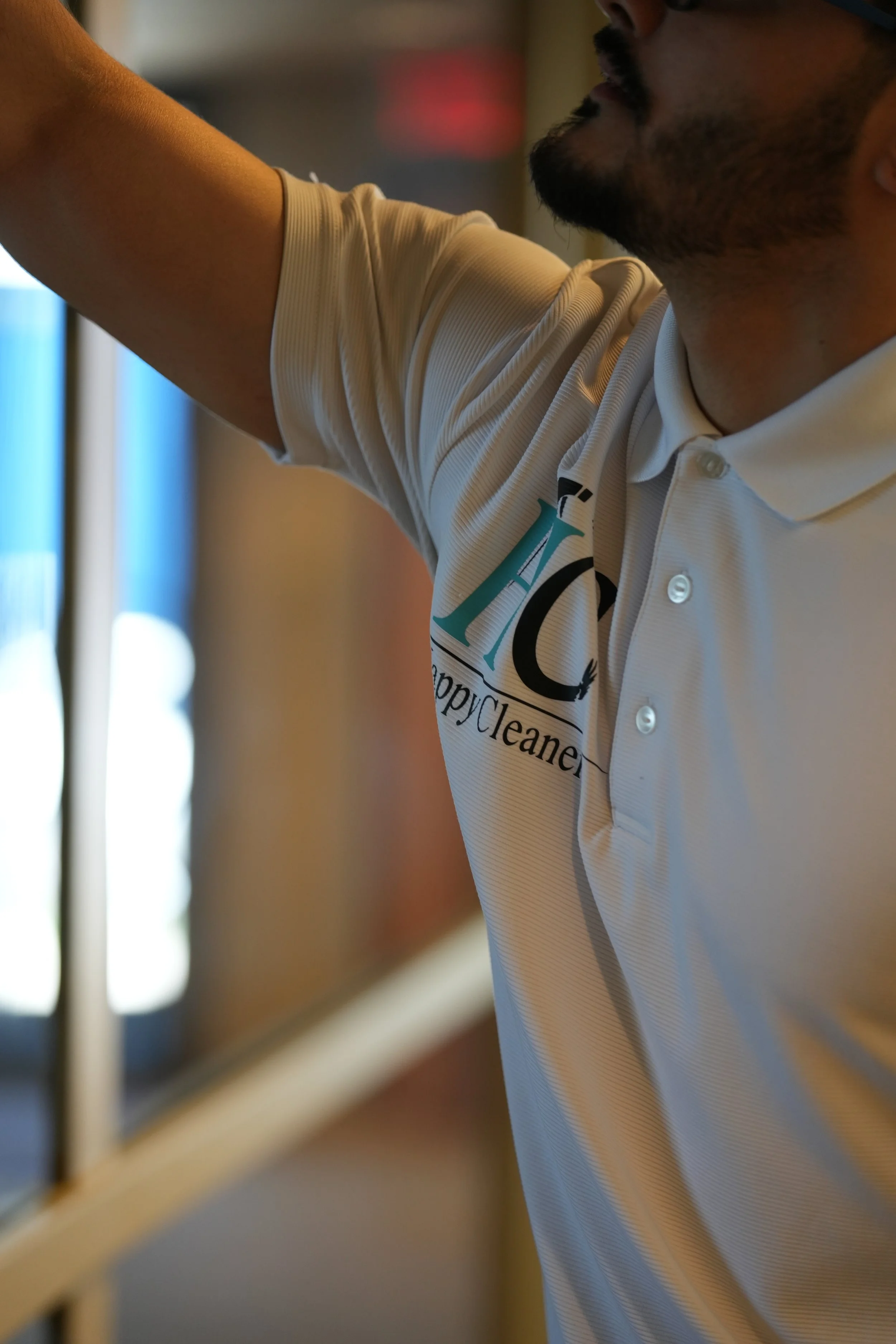 A person wearing a white polo shirt with a logo for a cleaning company, reaching towards a mirror, in an indoor setting.