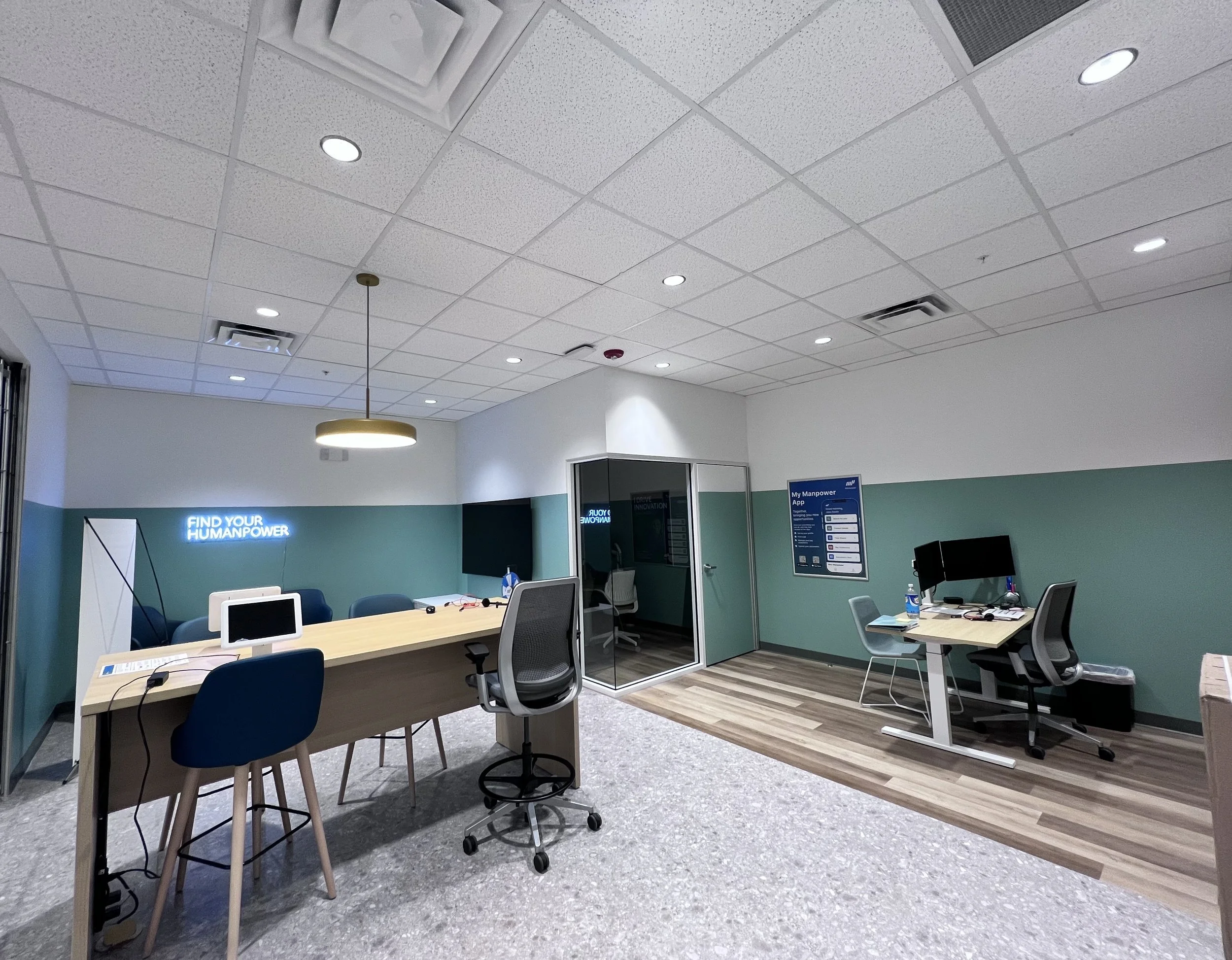 Office space with a long table, chairs, computers, a wall sign that reads 'Find Your Humanpower', a glass-fronted room, and a blue and white information poster on the wall.