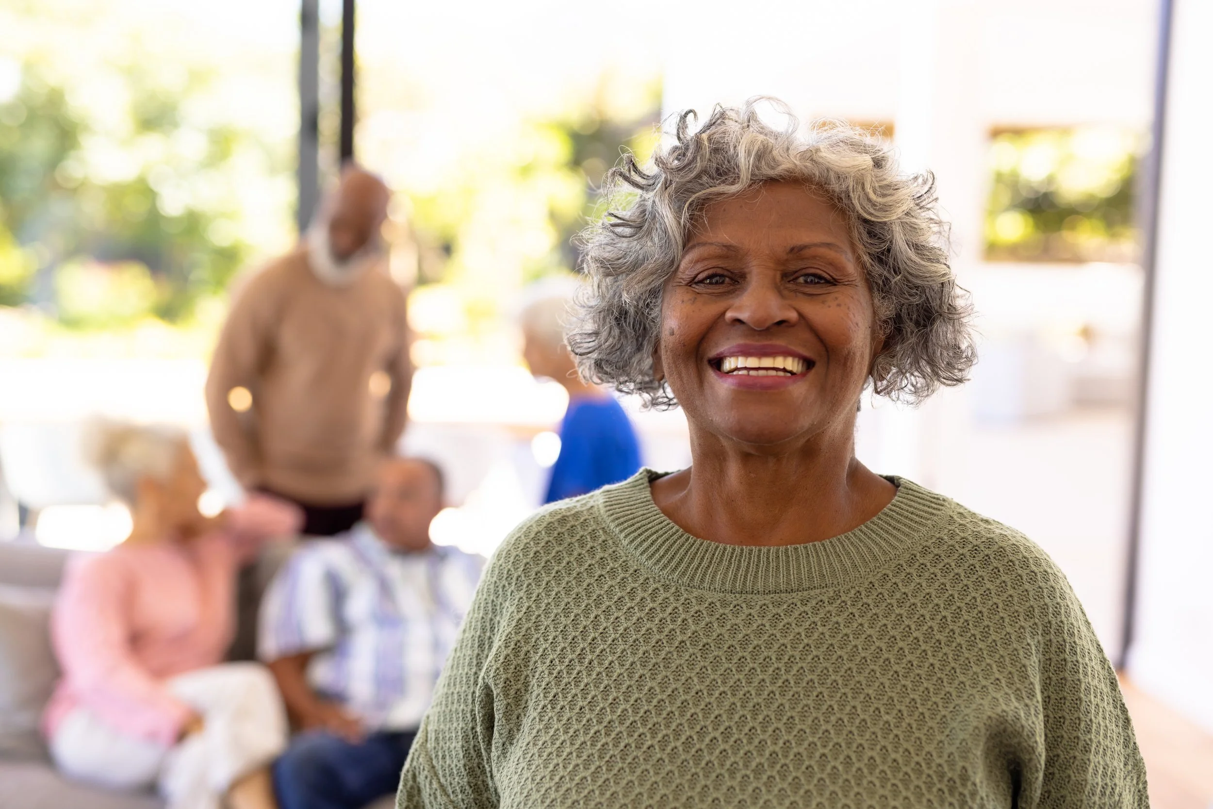 Smiling older African American woman in green sweater in a bright room with senior friends in the background.
