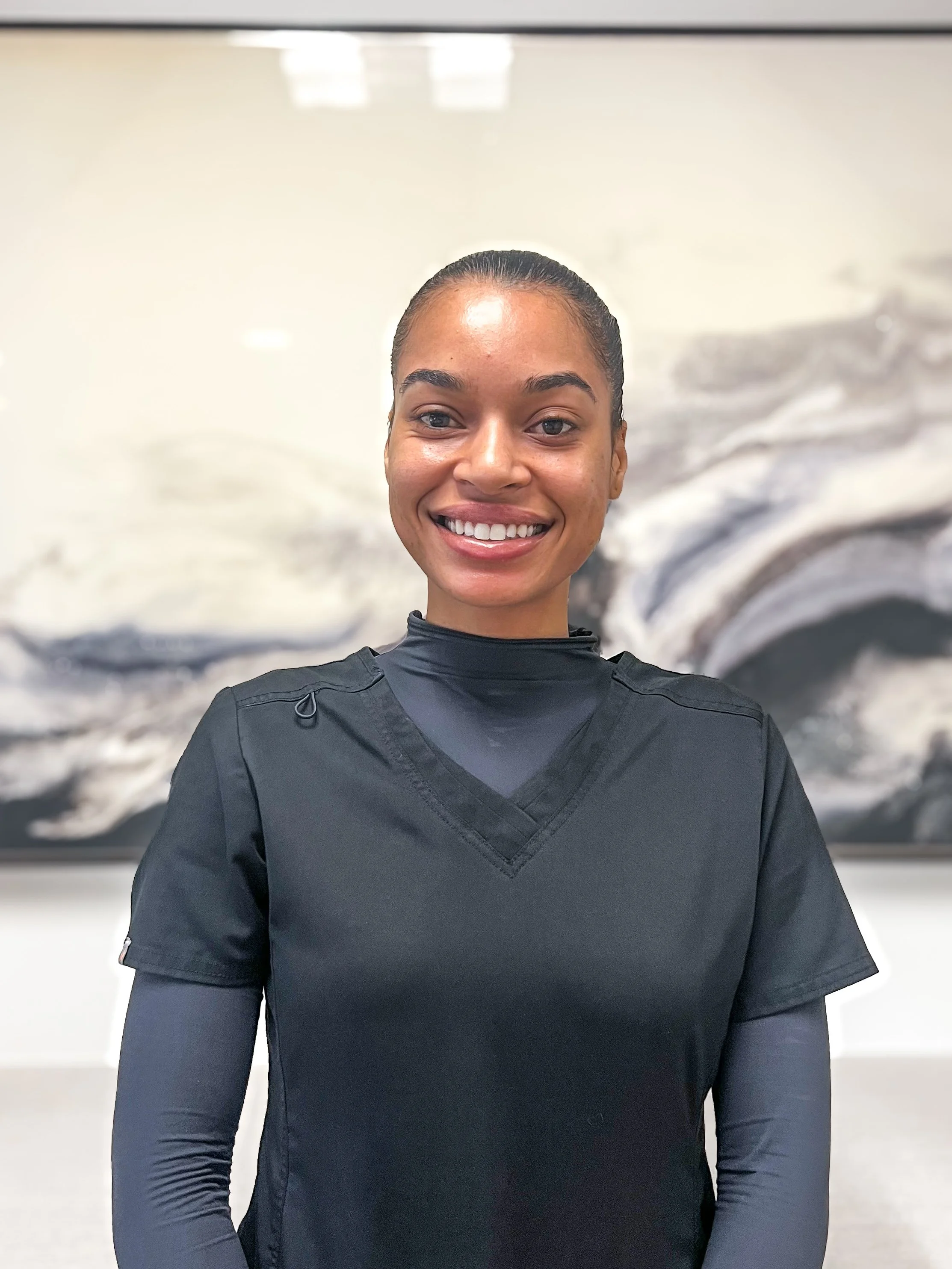 A woman with shoulder-length brown hair wearing a black medical scrub top, standing indoors with a blurred abstract painting in the background.