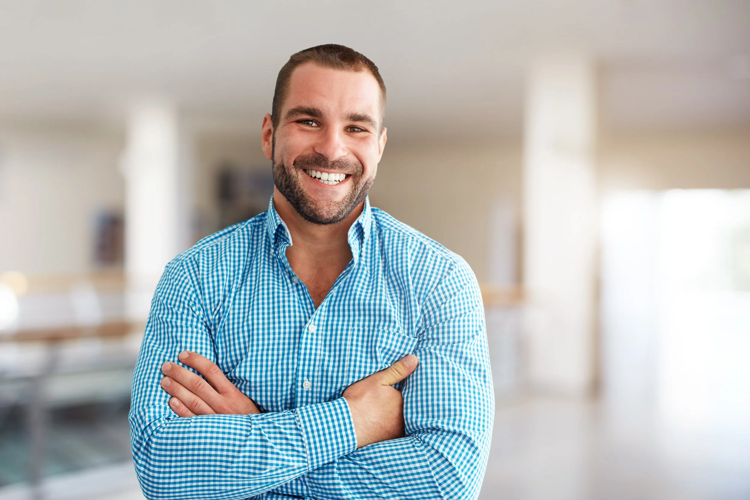 A smiling man with a beard and short hair, wearing a blue checkered shirt, standing with arms crossed in a bright, blurred indoor setting.