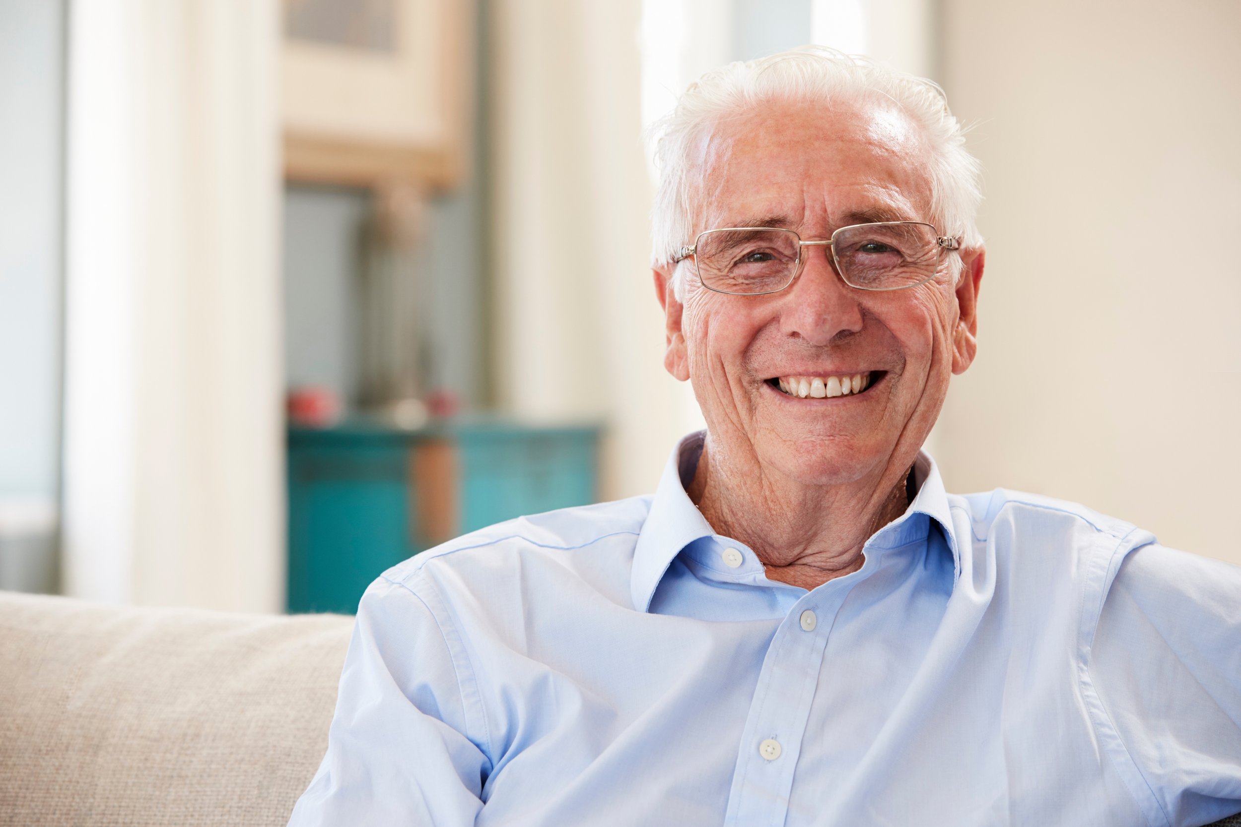 Smiling elderly man with white hair and glasses, wearing a light blue shirt, sitting on a beige sofa in a bright room.