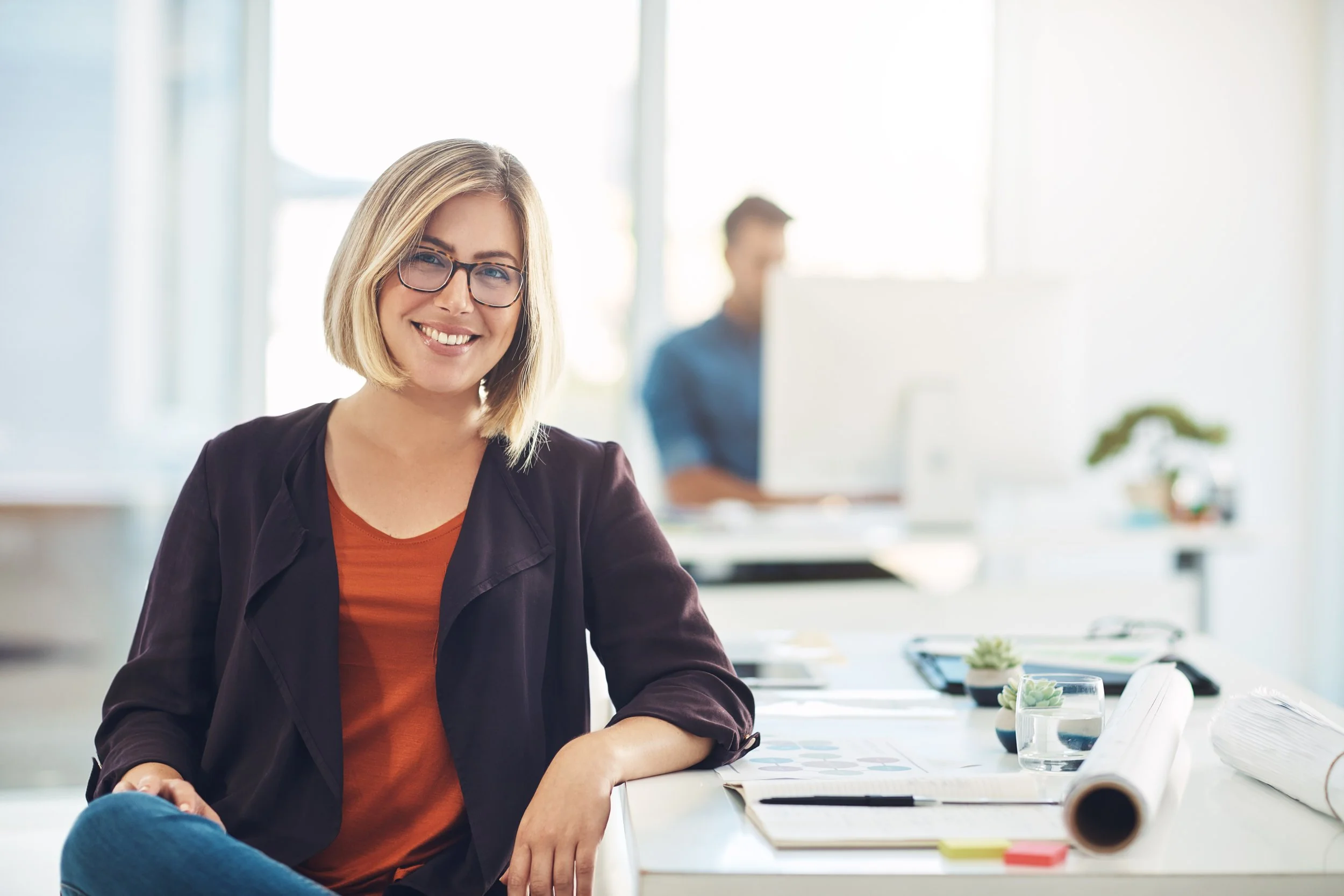 A woman with shoulder-length blonde hair and glasses smiling while sitting at her desk in an office, with a man working on a computer in the background.