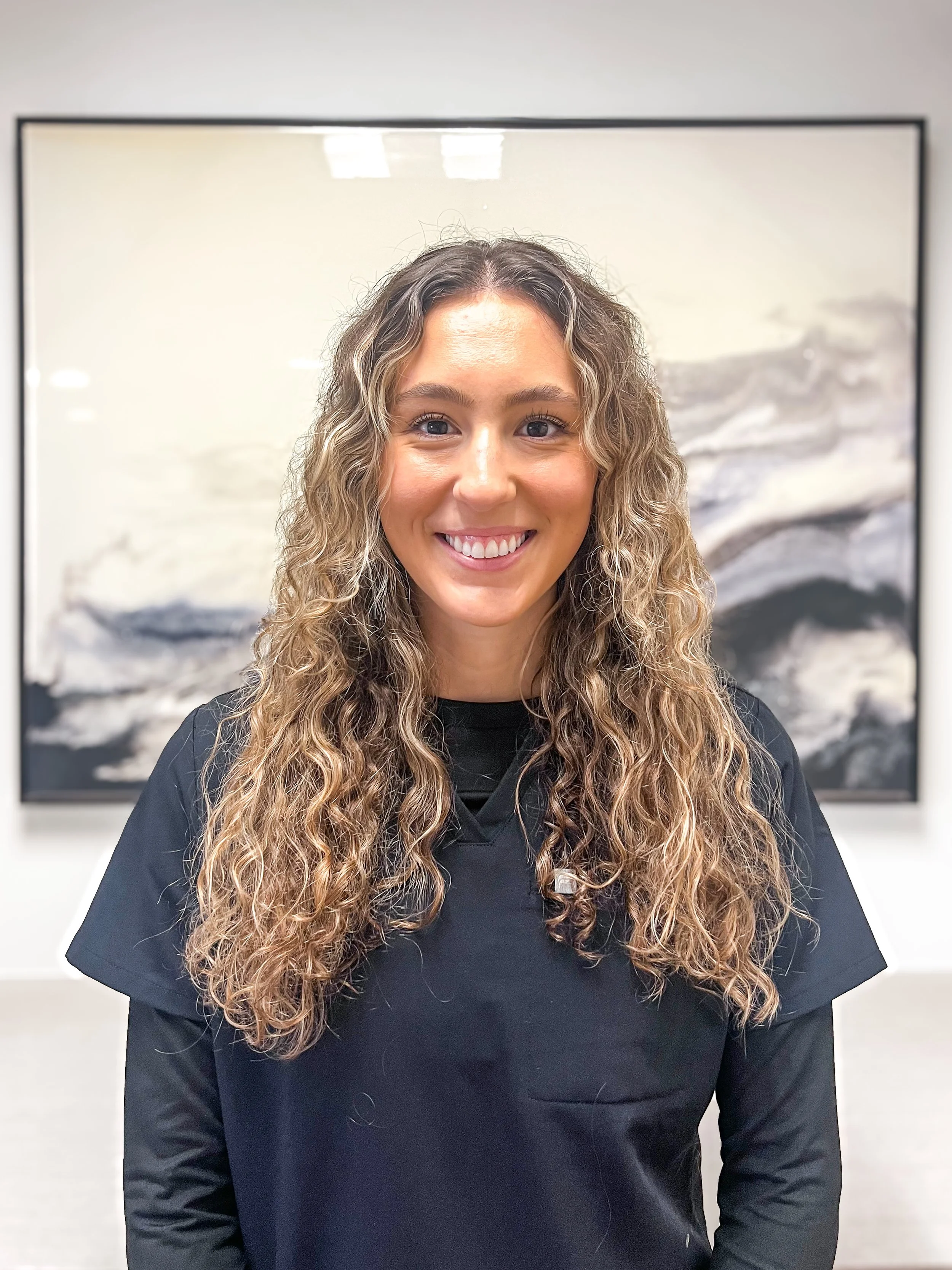 Young woman with long wavy hair and a nose ring smiling, wearing dark scrubs with a white long sleeve shirt underneath, standing indoors with an abstract black and white painting in the background.