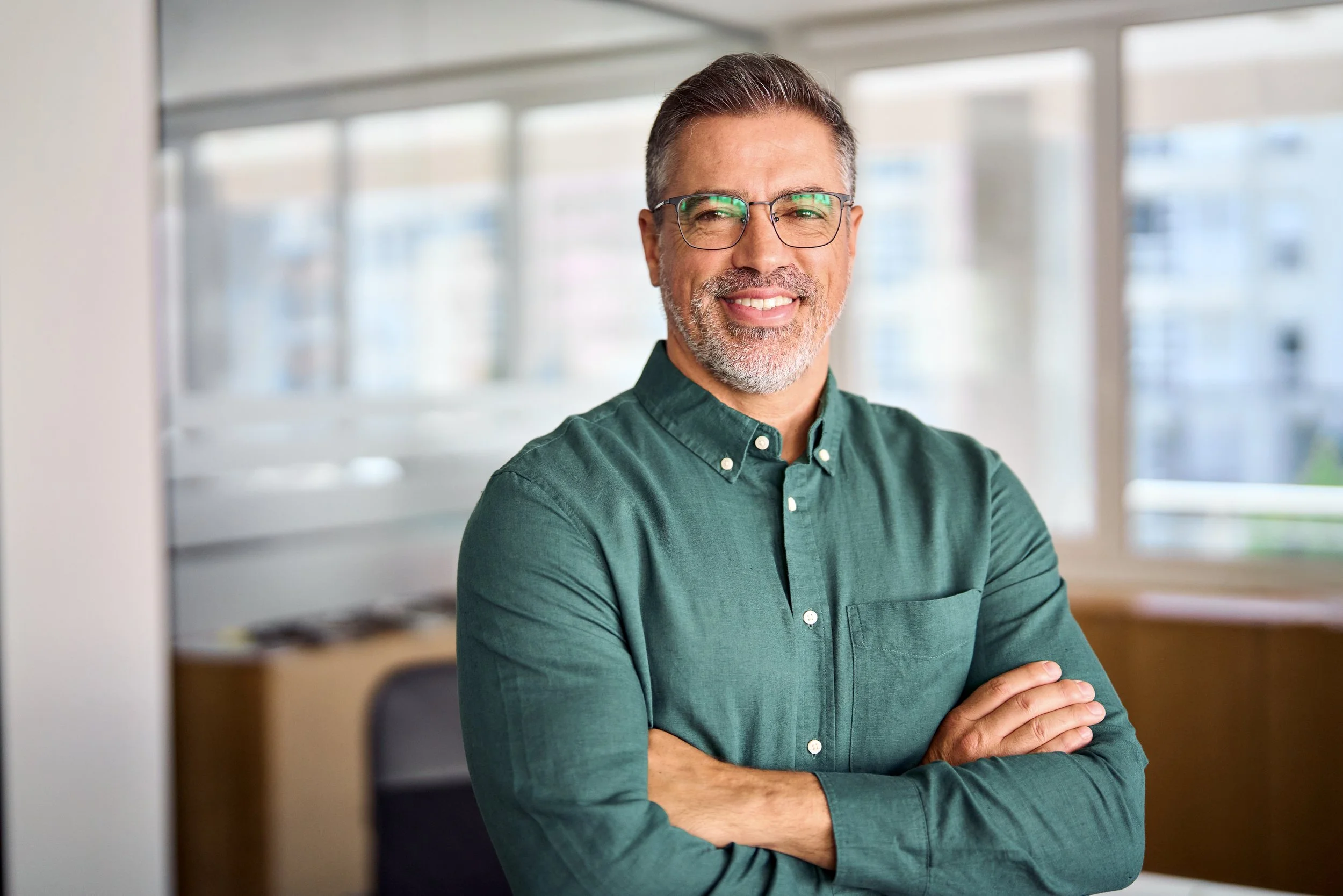 A smiling middle-aged man with glasses and gray hair, wearing a green button-up shirt, standing with arms crossed in a bright office with large windows.
