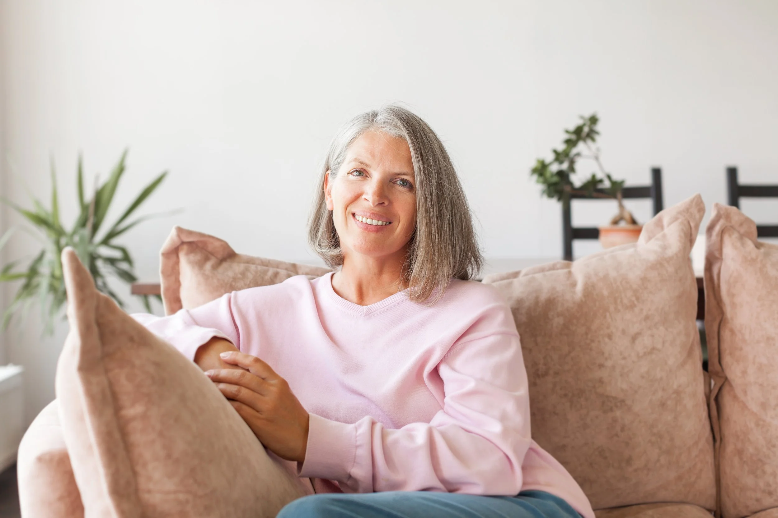 A mature woman with gray hair wearing a pink sweater sitting on a beige couch with cushions, smiling at the camera in a bright living room with a potted plant in the background.