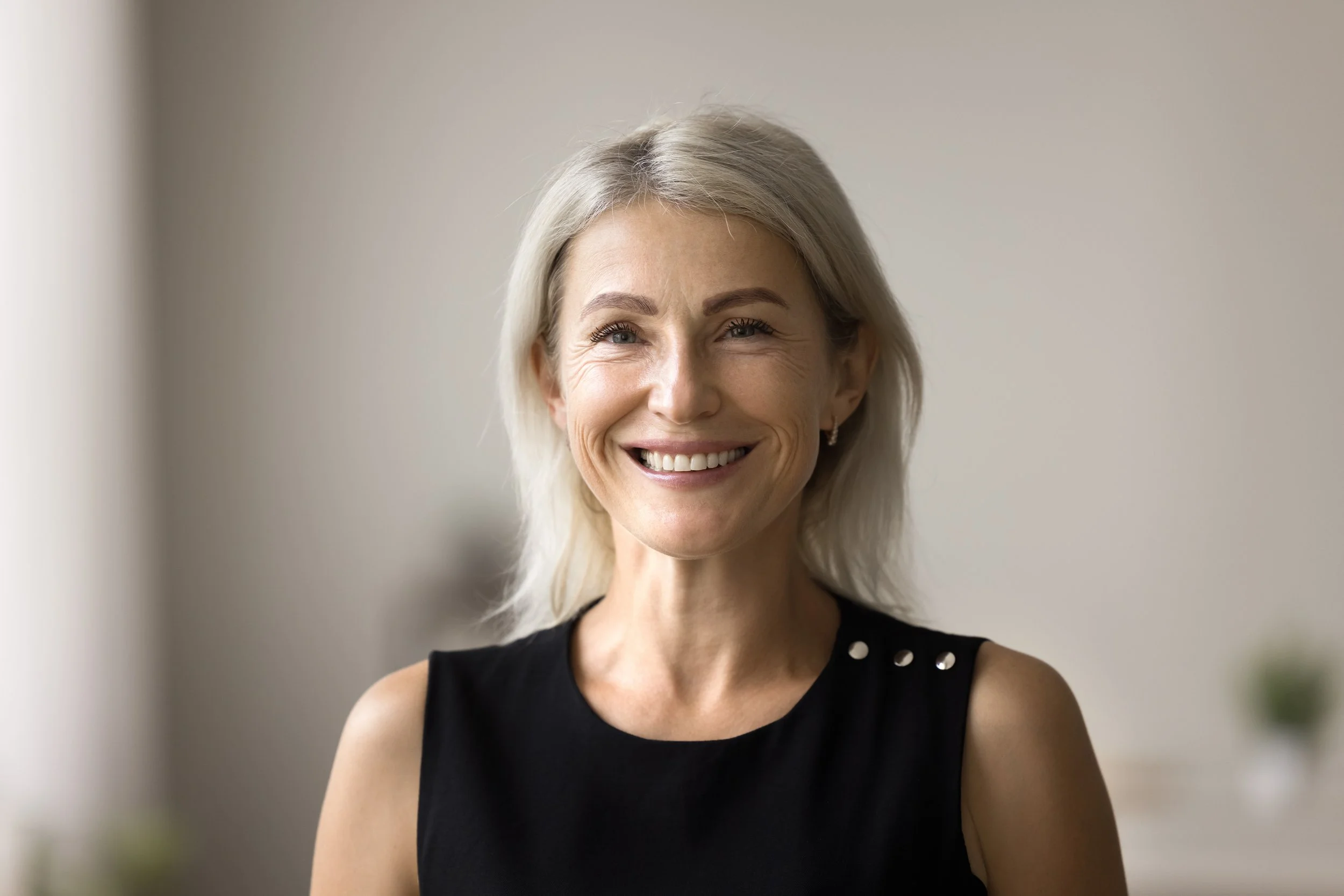 Smiling older woman with shoulder-length gray hair wearing a black sleeveless top with metal studs on the shoulder, standing indoors with a neutral background.