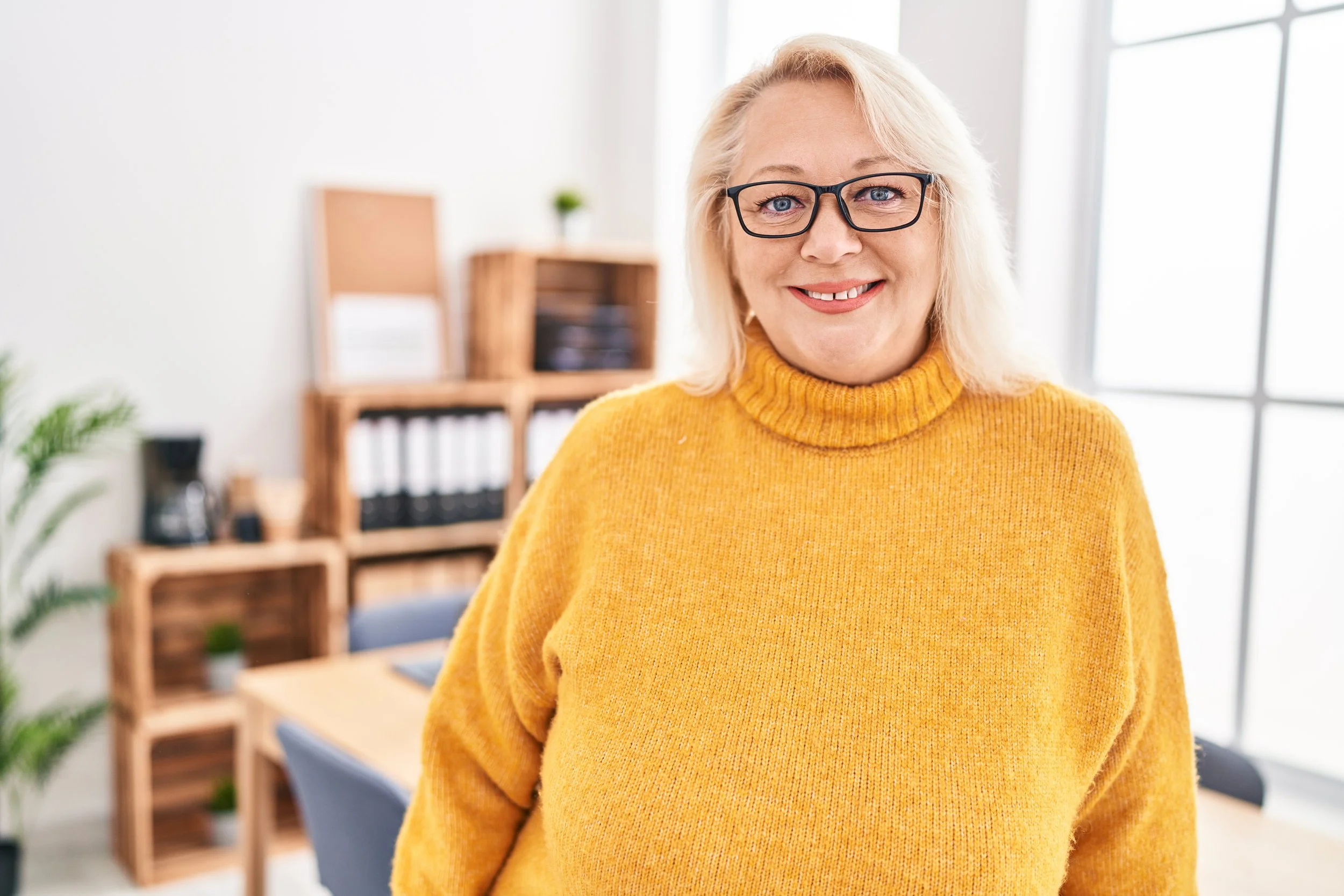 Smiling woman with blonde hair, wearing glasses and a yellow sweater, standing in a bright, modern office with wooden shelves and a large window.