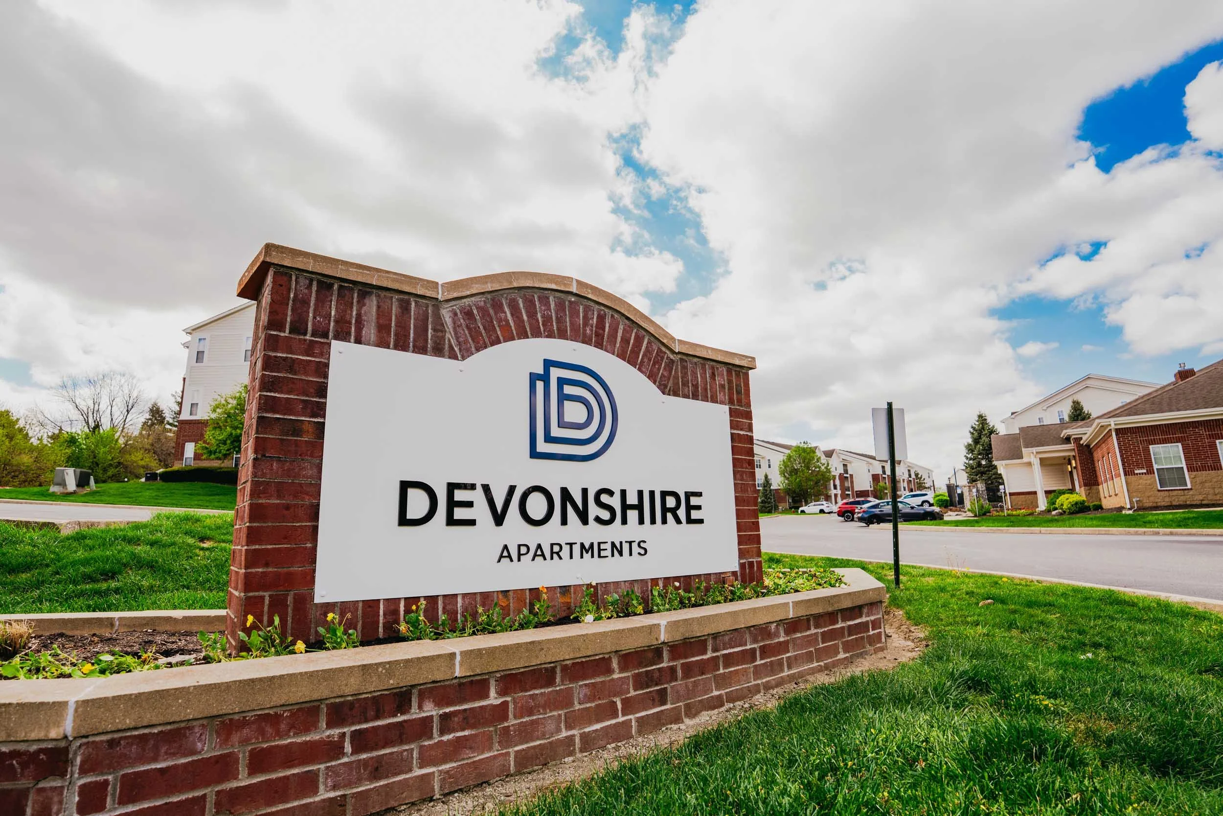 Sign for Devonshire Apartments on a brick structure with a city street and residential buildings in the background under a cloudy sky.
