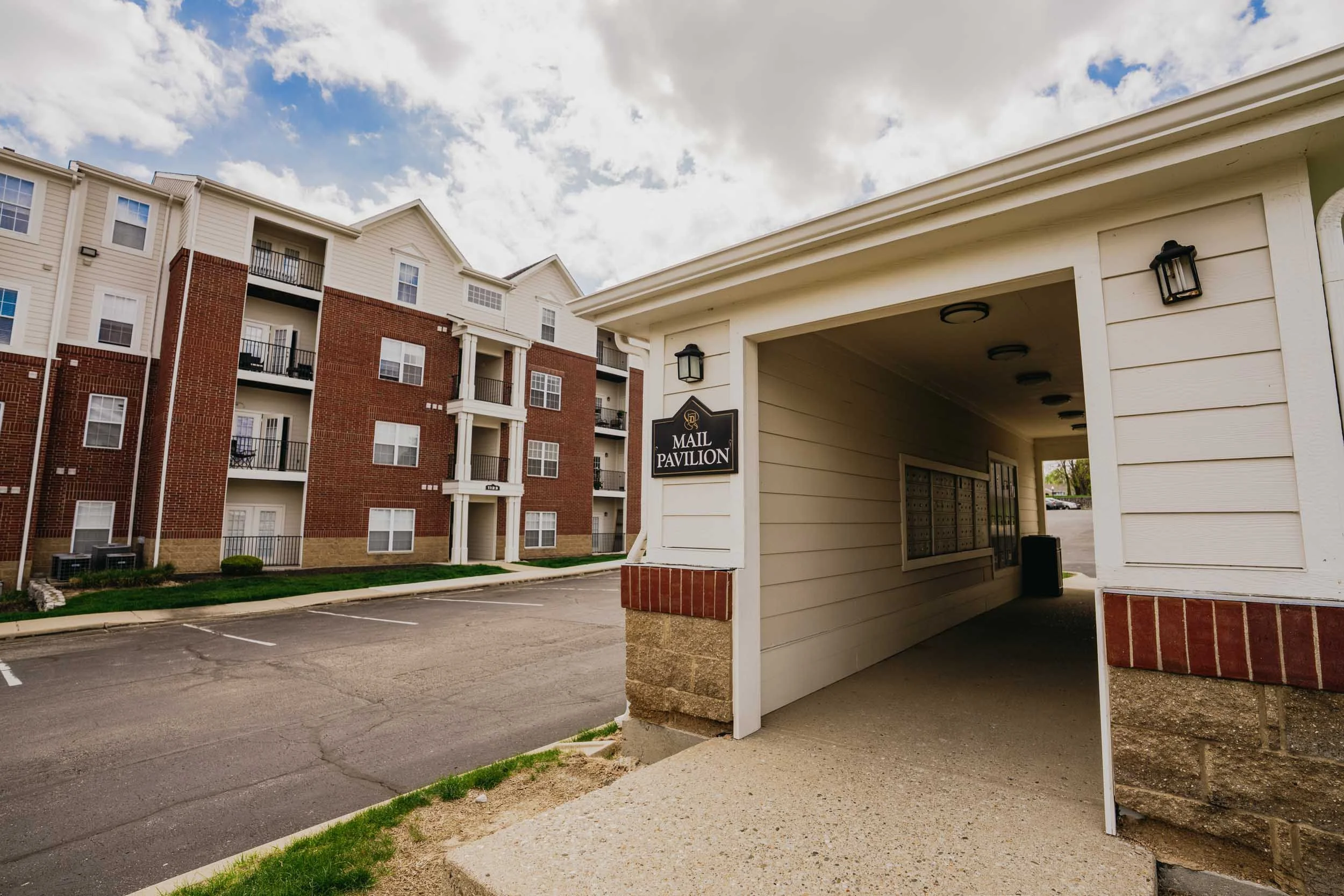 Apartment complex with red brick and beige siding building, mail pavilion with multiple mailboxes, parking lot, and partly cloudy sky.