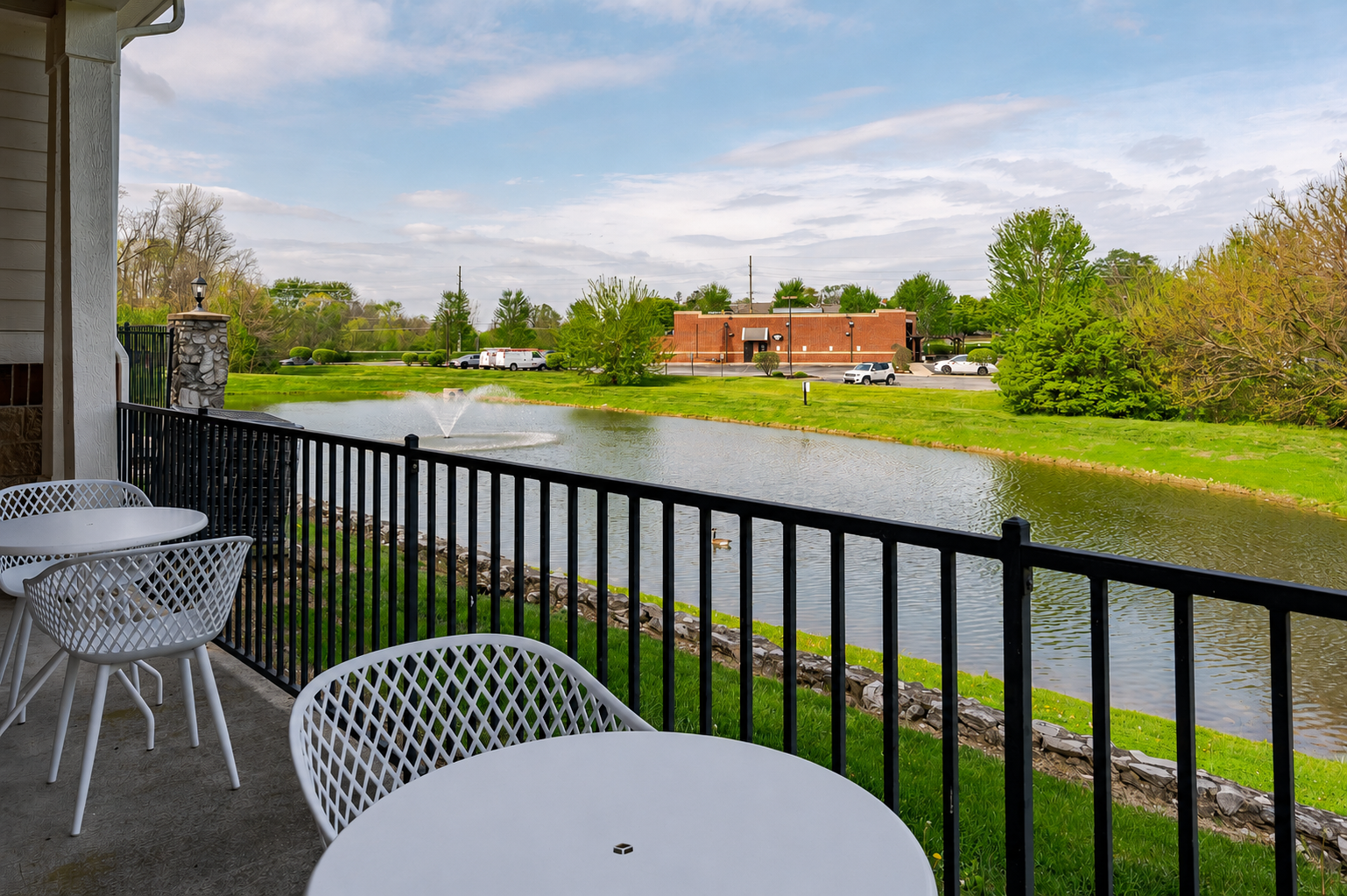 Patio with white tables and chairs overlooking a pond with a fountain, surrounded by green grass and trees.