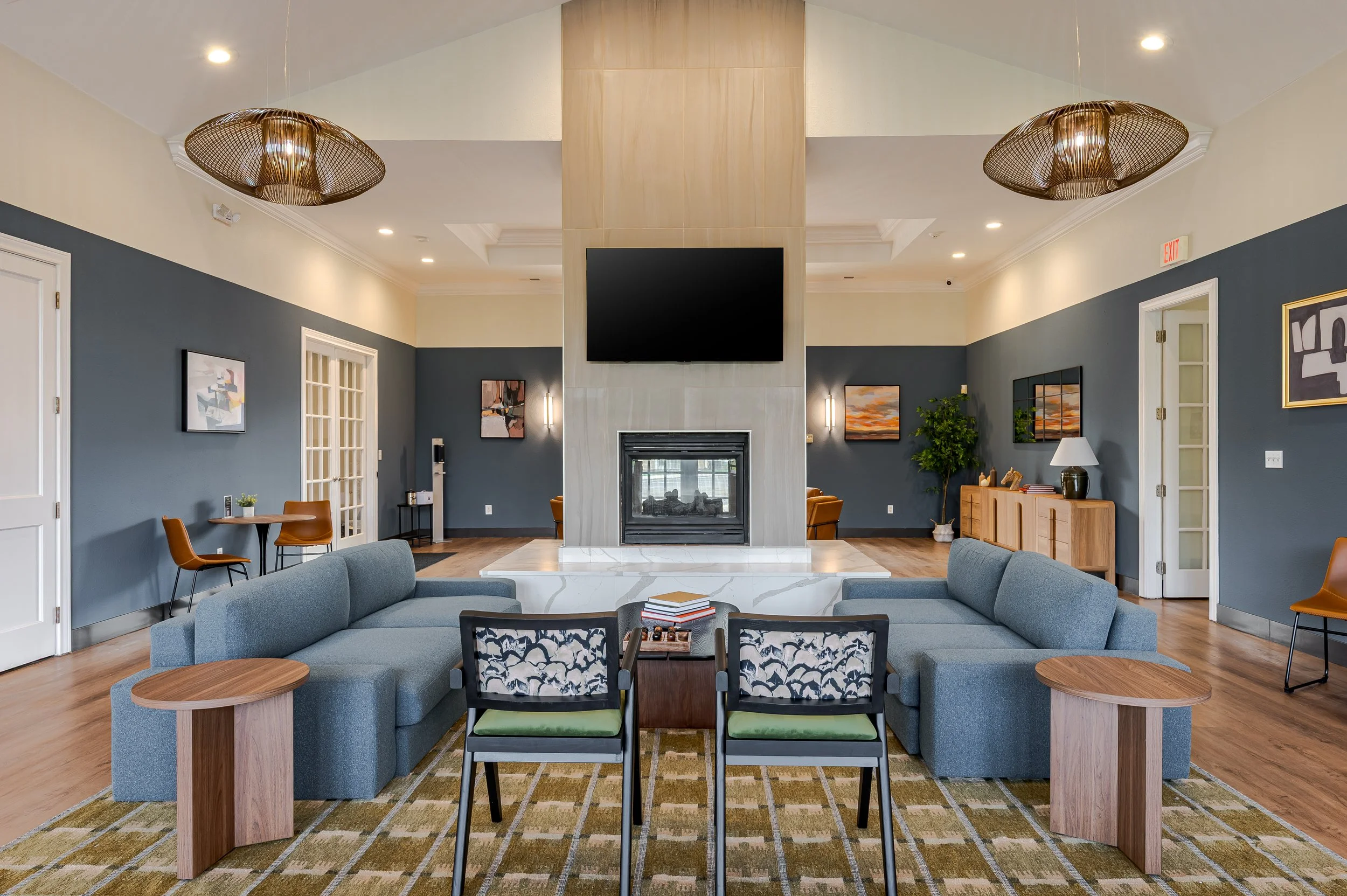 A spacious living room with gray couches, a fireplace, wall-mounted TV, artwork, wooden side tables, and a patterned area rug.