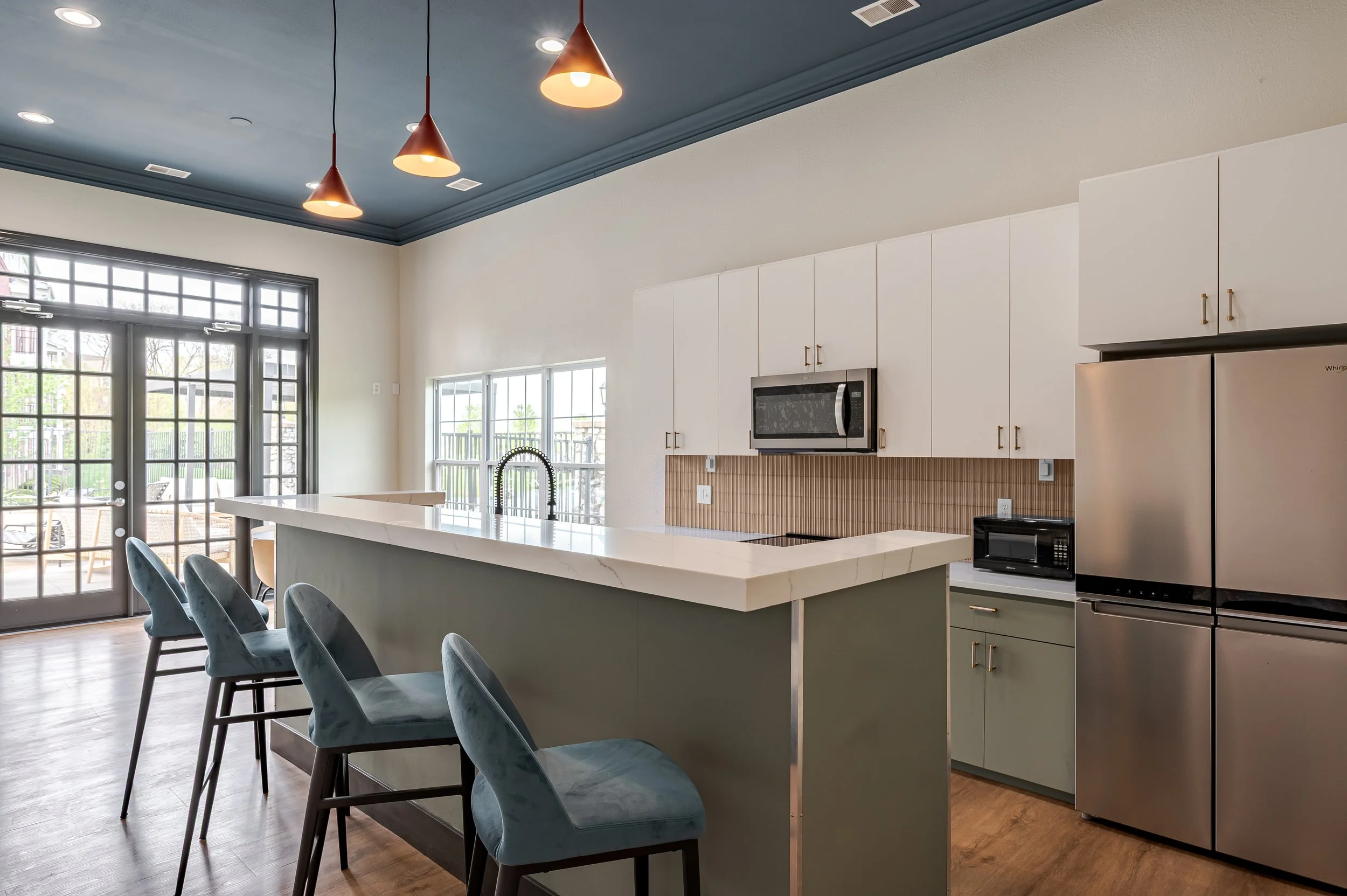 Modern kitchen with white cabinets, stainless steel appliances including a refrigerator, microwave, and oven, a large island with a white countertop, and four blue upholstered chairs. Large glass doors and windows let in natural light.