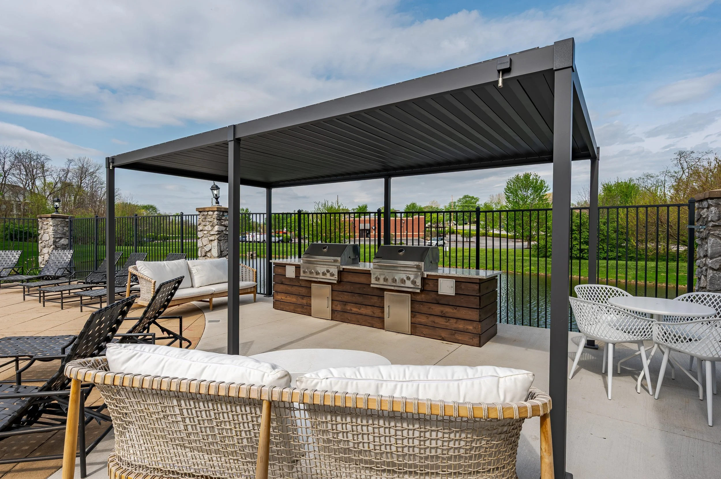 Outdoor patio with black chairs, a beige sofa, white round chairs, a built-in grill with a wooden cover, a metal fence, and a pergola with a cloudy sky in the background.