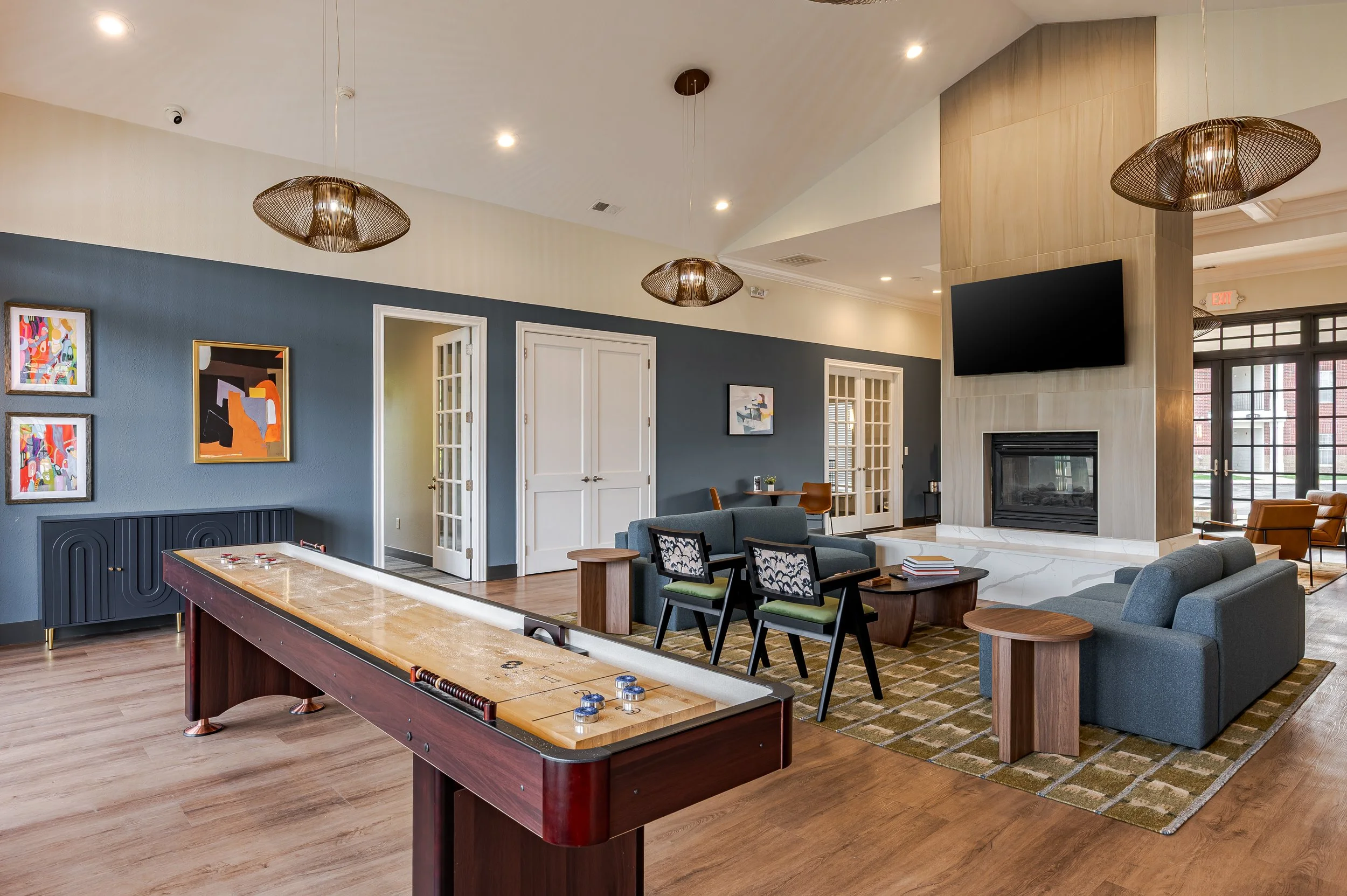 A spacious, modern lounge with artwork on a blue wall, a fireplace, a large flat-screen TV, a pool table, and comfortable seating including sofas and chairs. There are ceiling lights and large windows letting in natural light.